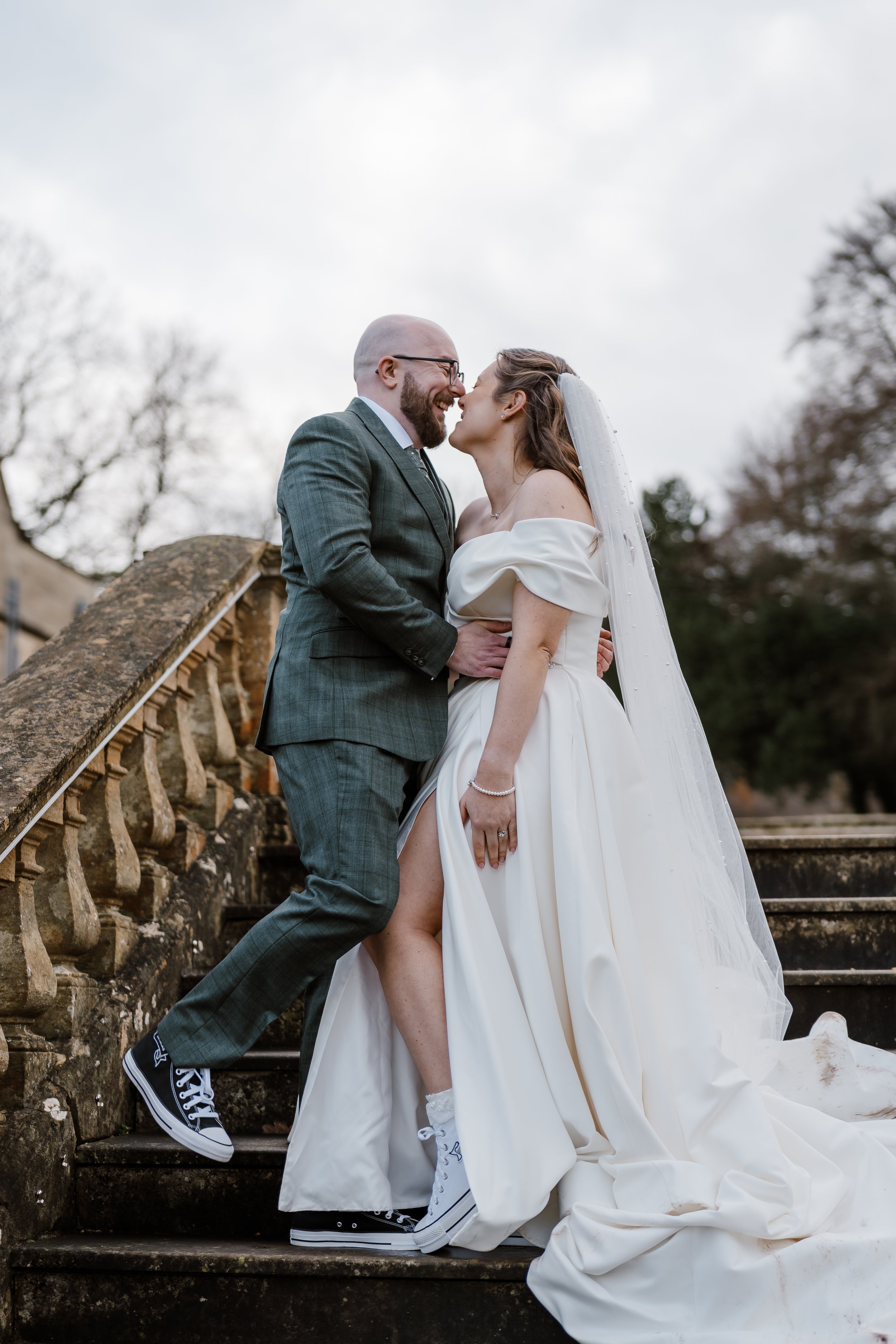 A newlywed couple dressed in wedding attire, wearing sneakers, standing on outdoor stone steps, shown smiling and touching noses.