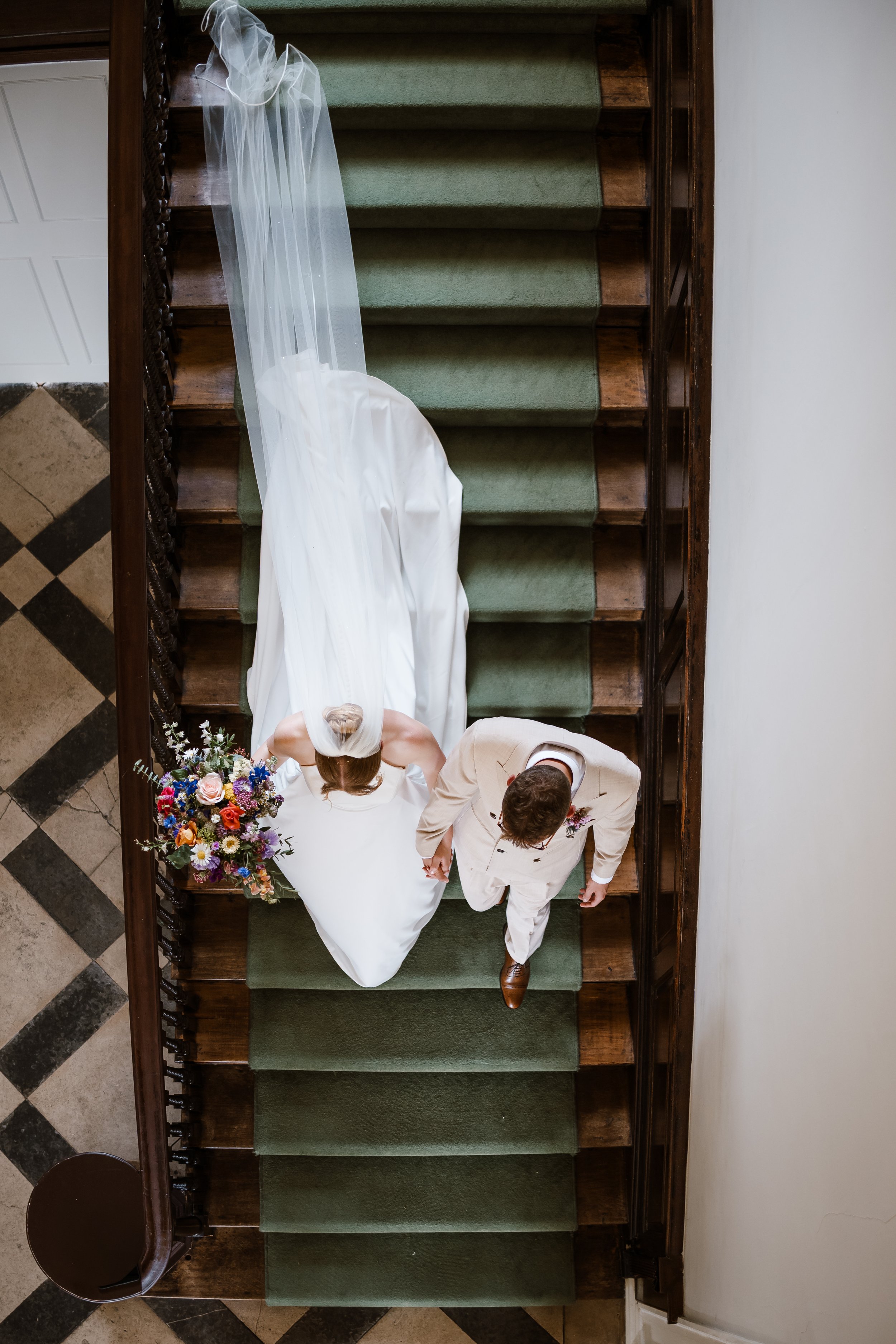 A bride wearing a white wedding dress and veil standing on a staircase, holding a bouquet of multicolored flowers, accompanied by a man in a beige suit.