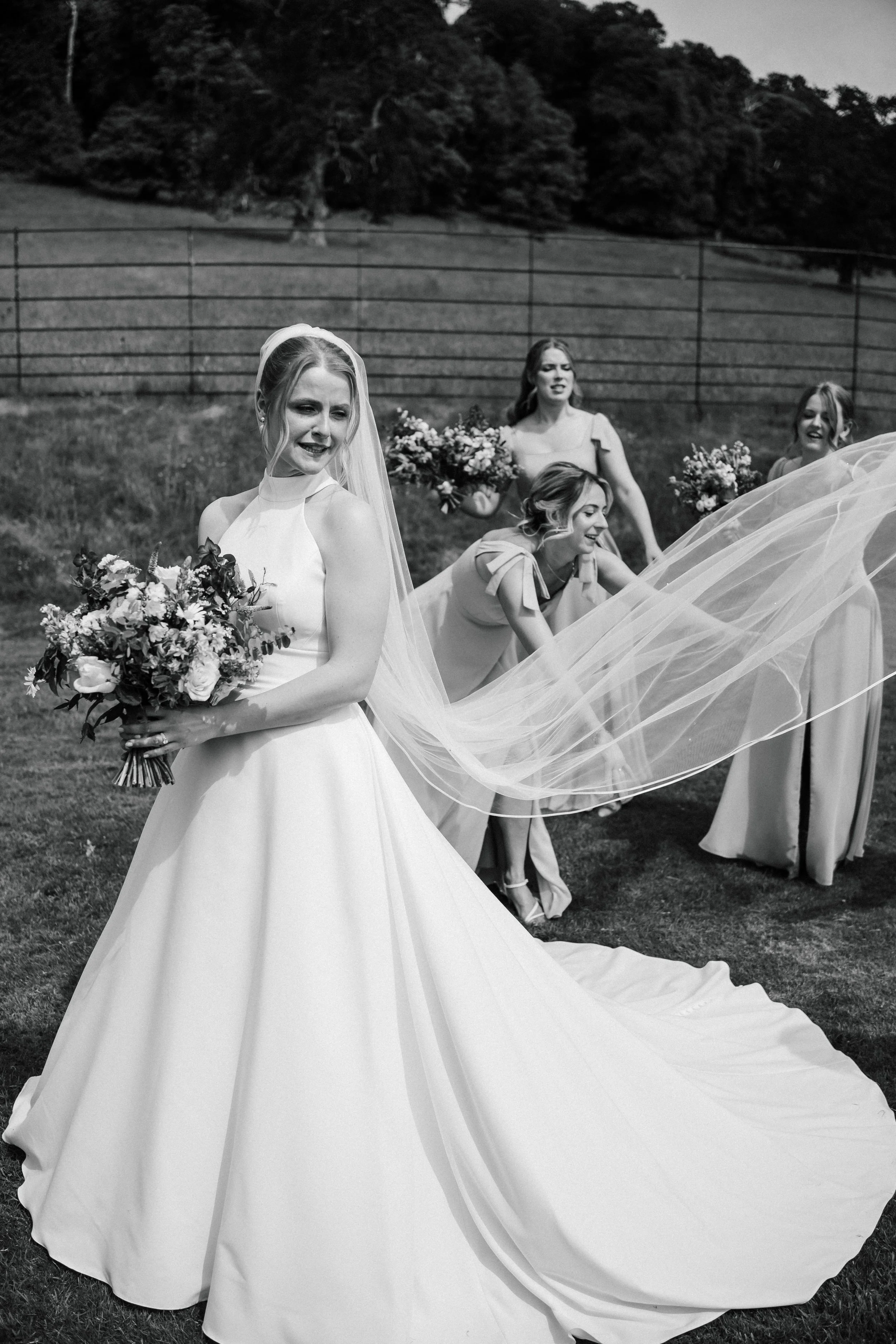 A bride in a wedding dress holding a bouquet of flowers, with bridesmaids in dresses holding bouquets in the background, outdoors on grass.