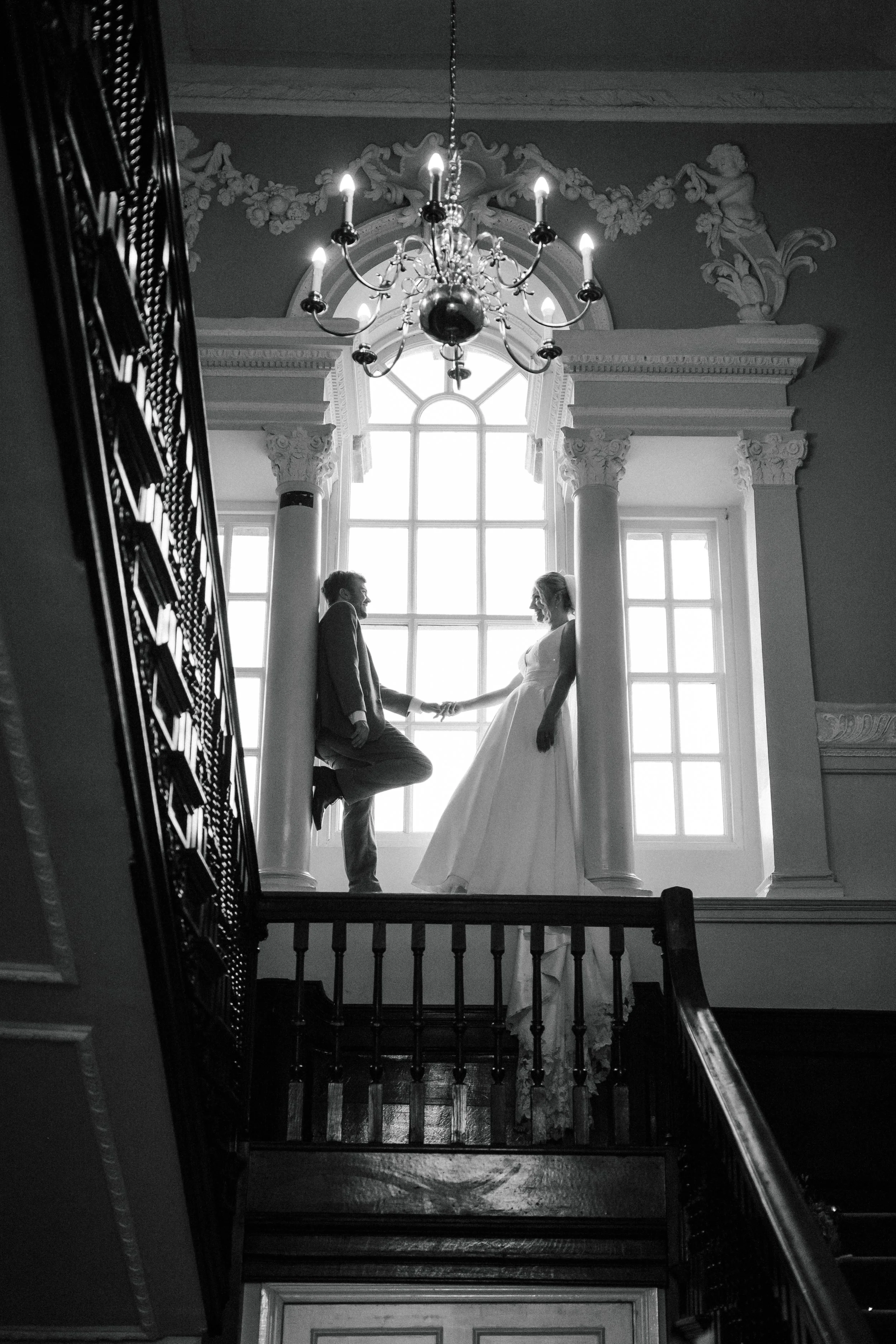Black and white photo of a couple in wedding attire holding hands, standing on a balcony with large windows and ornate columns, with a chandelier hanging above.