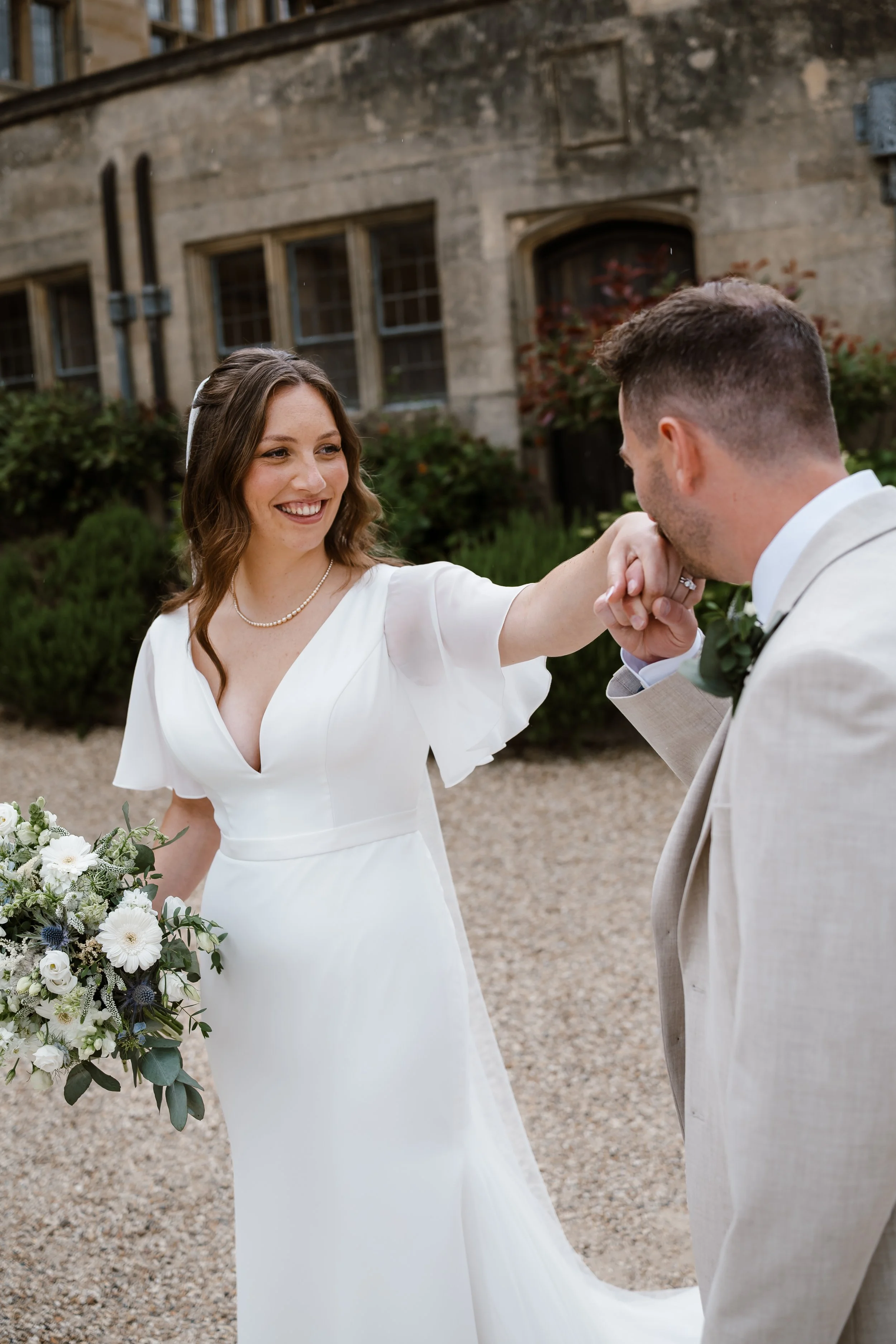 A bride and groom are holding hands in an outdoor setting during their wedding ceremony. The bride is smiling, wearing a white wedding dress and a pearl necklace, with brown hair styled in loose waves. The groom, dressed in a light-colored suit, is k