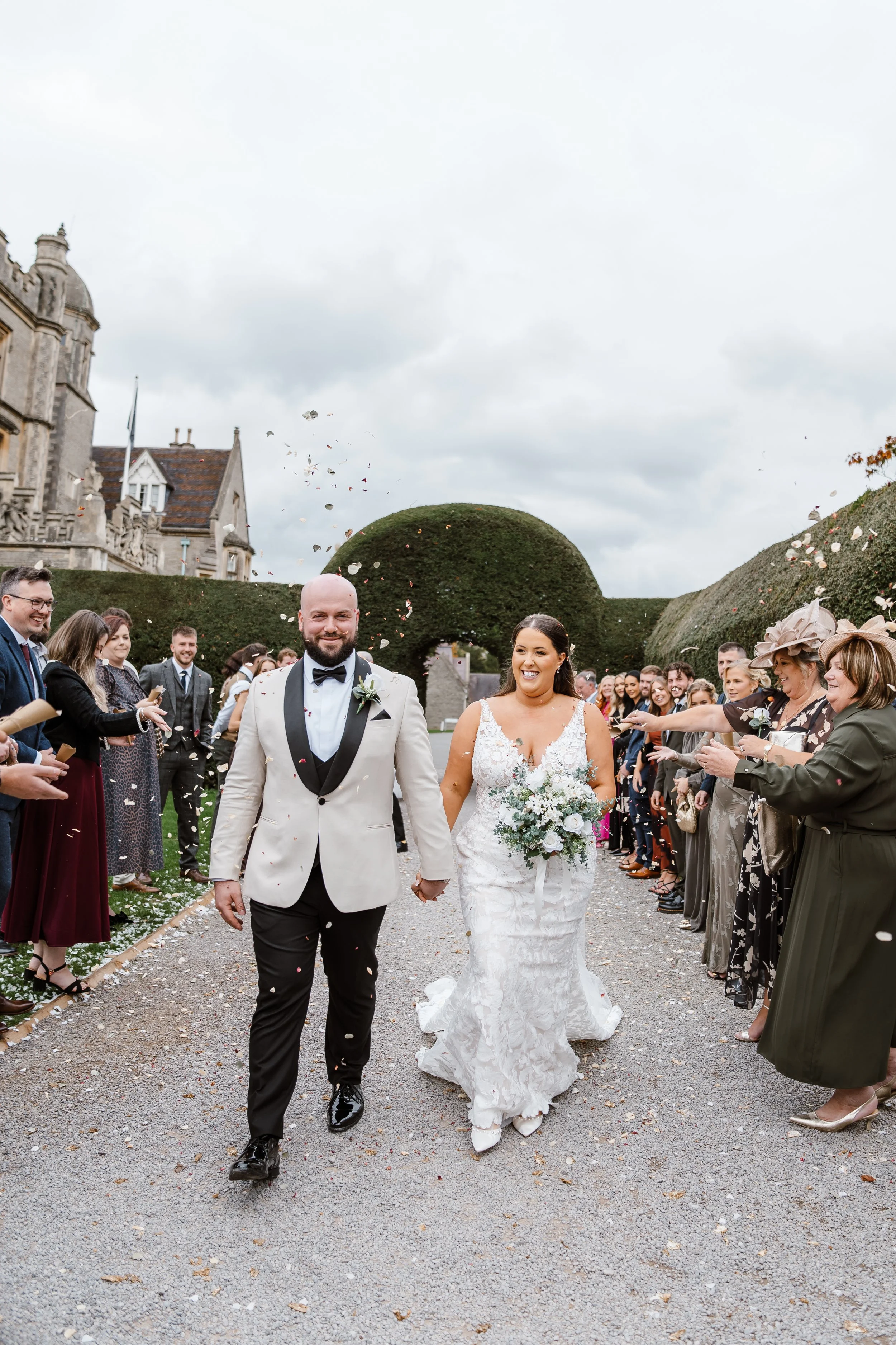 A newlywed couple walks hand in hand down a wedding aisle, smiling, as guests throw confetti in the air during an outdoor wedding ceremony on a cloudy day.