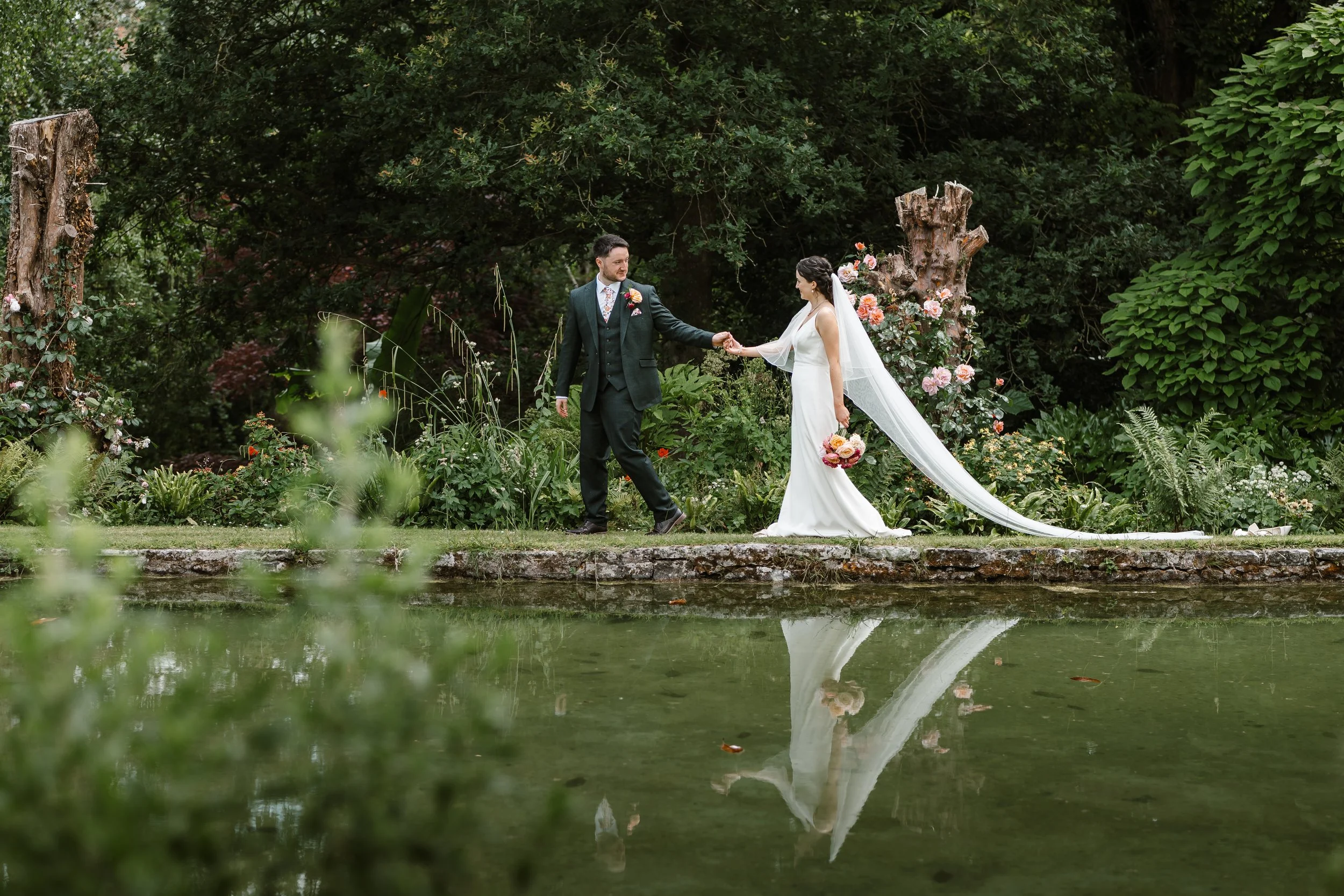 A bride and groom holding hands in a lush green garden by a pond, with the bride in a white wedding dress and veil, and the groom in a dark suit, surrounded by trees and flowering plants.