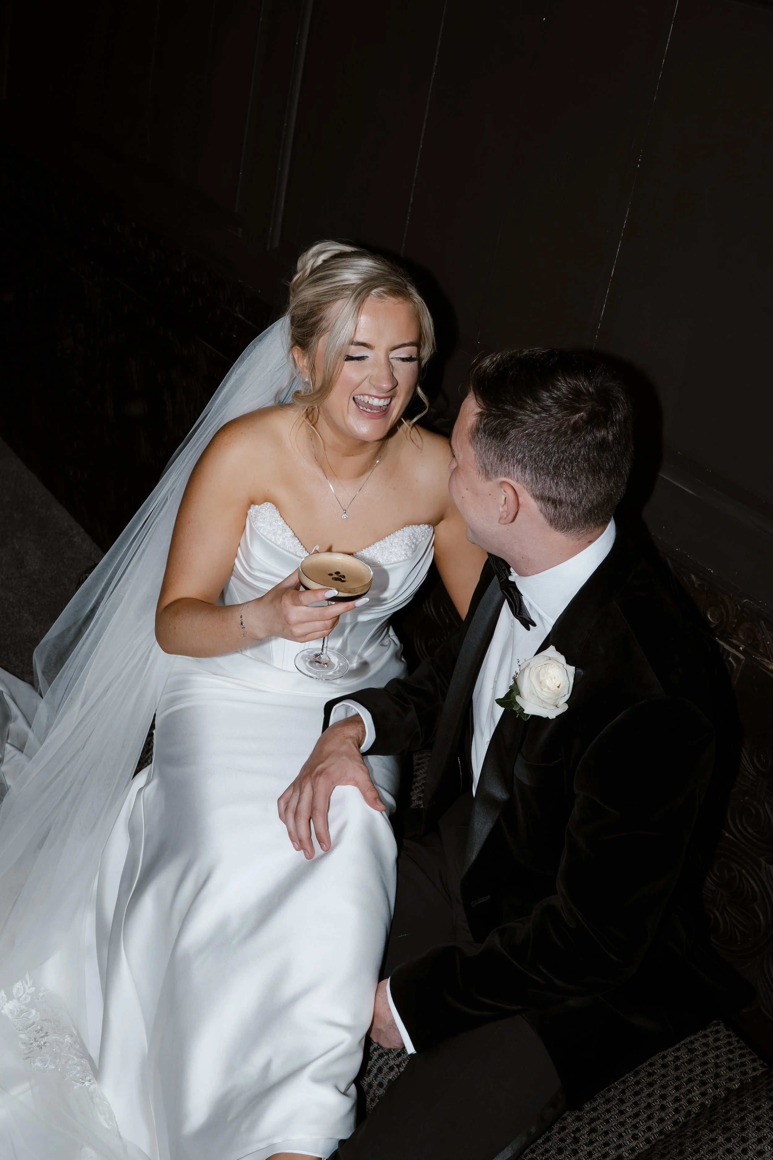 A bride in a white wedding dress and veil smiling and laughing while holding a glass of champagne, sitting next to a groom in a black tuxedo with a white boutonniere, both looking at each other and sharing a joyful moment.