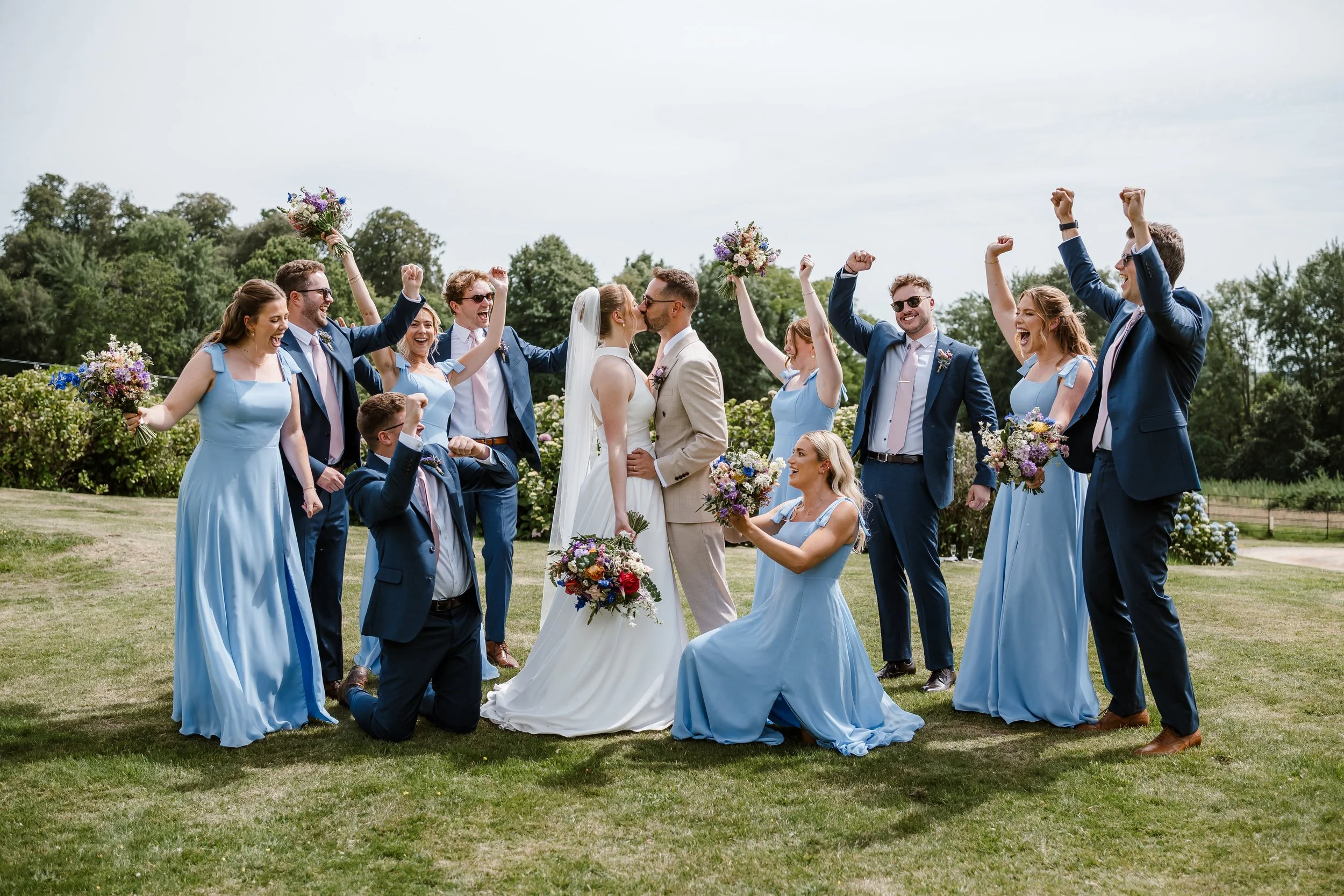 A group of wedding guests, dressed in blue formal wear, celebrate outdoors during a wedding ceremony. The bride and groom kiss in the center, while the others cheer and raise their fists in excitement, holding bouquets of flowers.