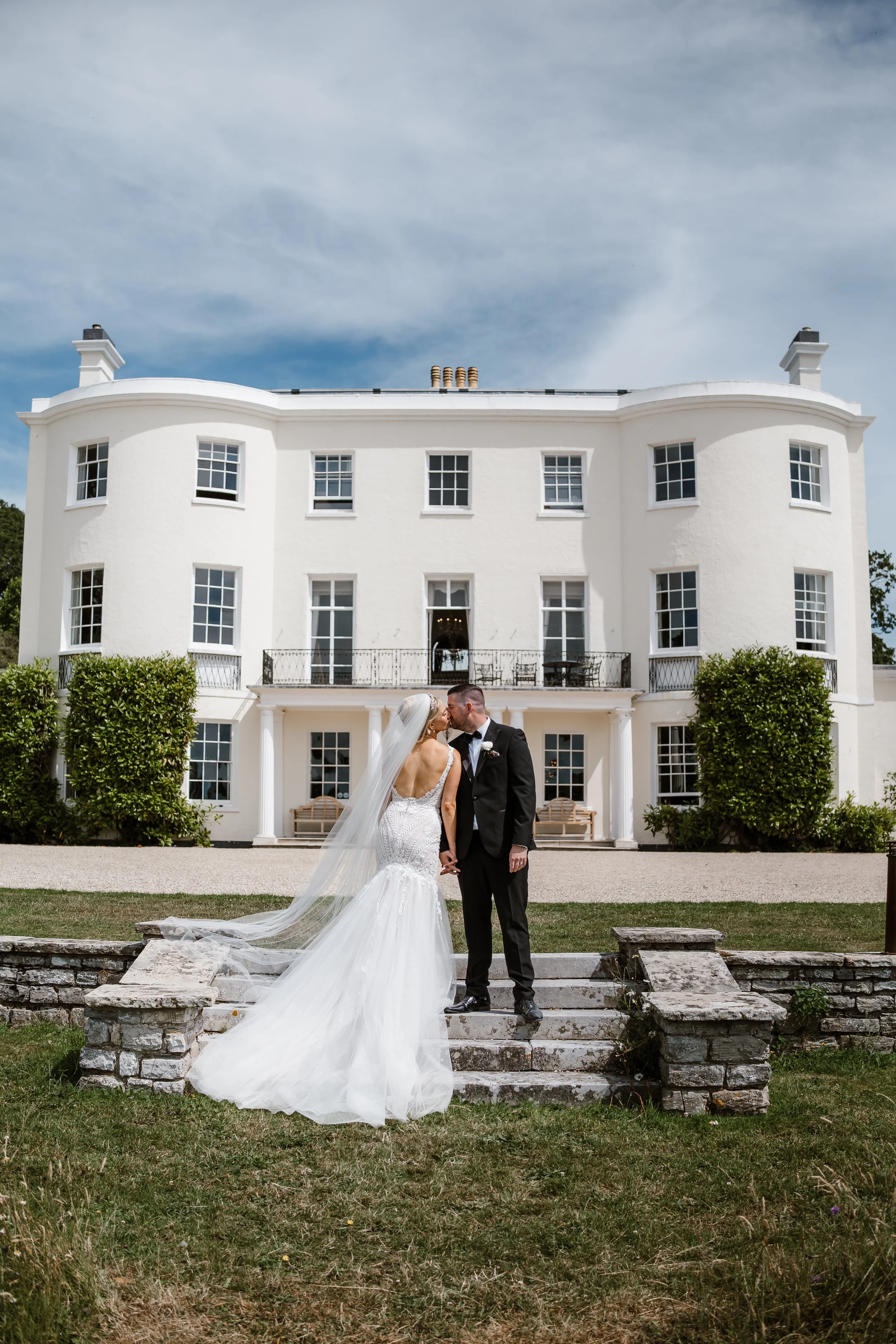 A bride and groom kissing on stone steps in front of a large white mansion during a sunny day, with the bride wearing a long train and veil, and the groom in a black tuxedo.