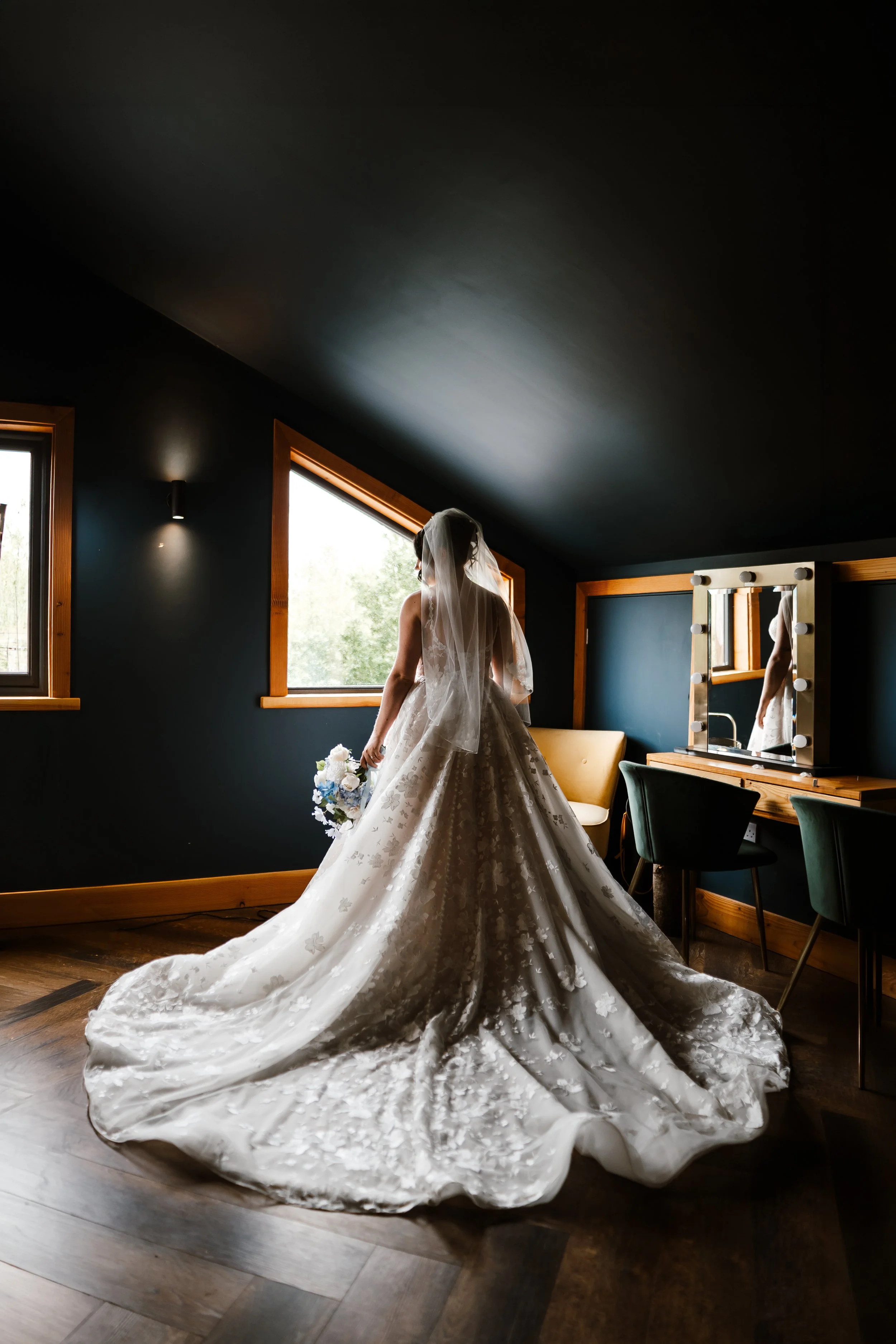 A bride in a wedding dress with a long train standing in a dark room, gazing out of a window, holding a bouquet of flowers.