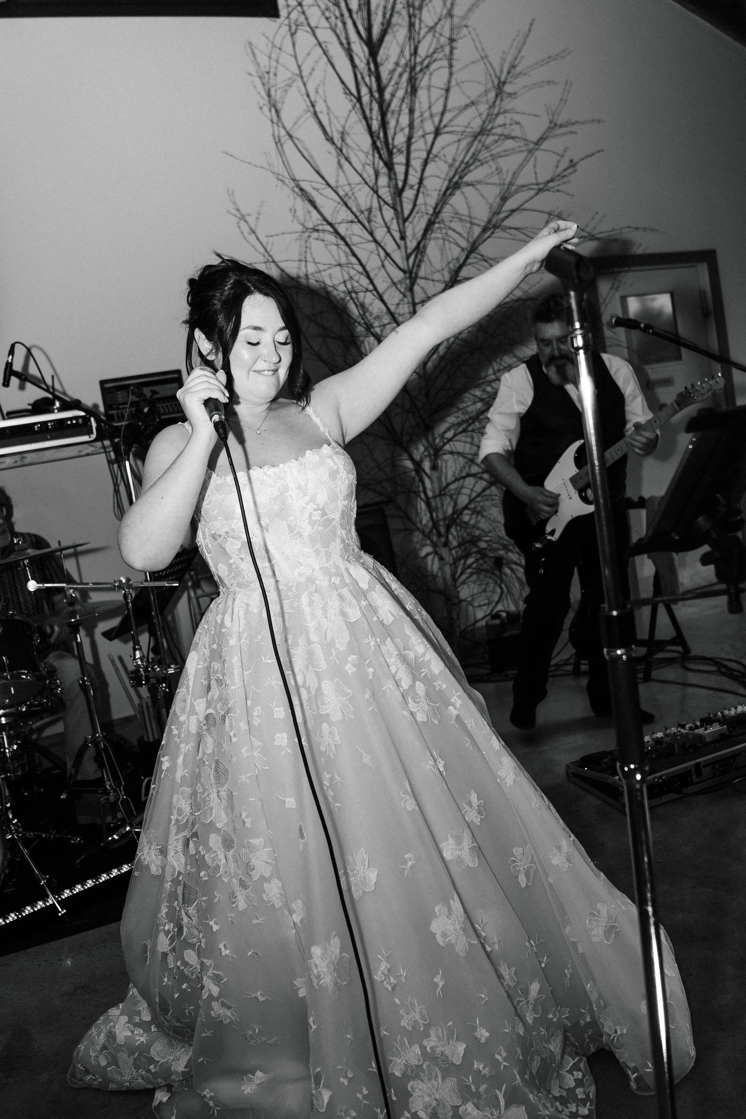 A woman in a strapless, floral ball gown singing into a microphone on stage. A man playing an electric guitar is in the background, with musical equipment and a tree without leaves behind them.