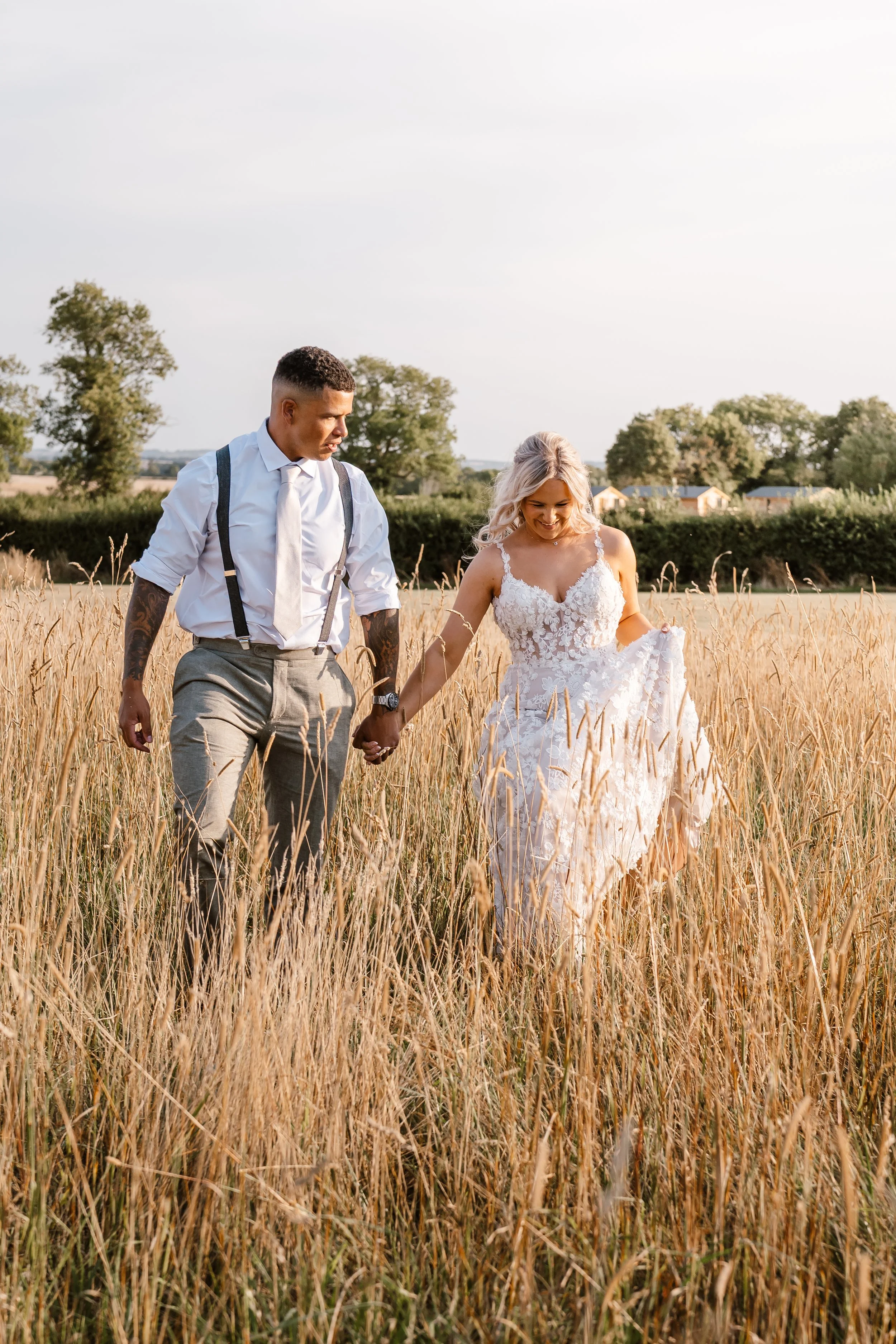 A newlywed couple walking hand in hand through a golden wheat field, the bride in a white lace wedding dress and the groom in a white shirt with suspenders, during sunset.