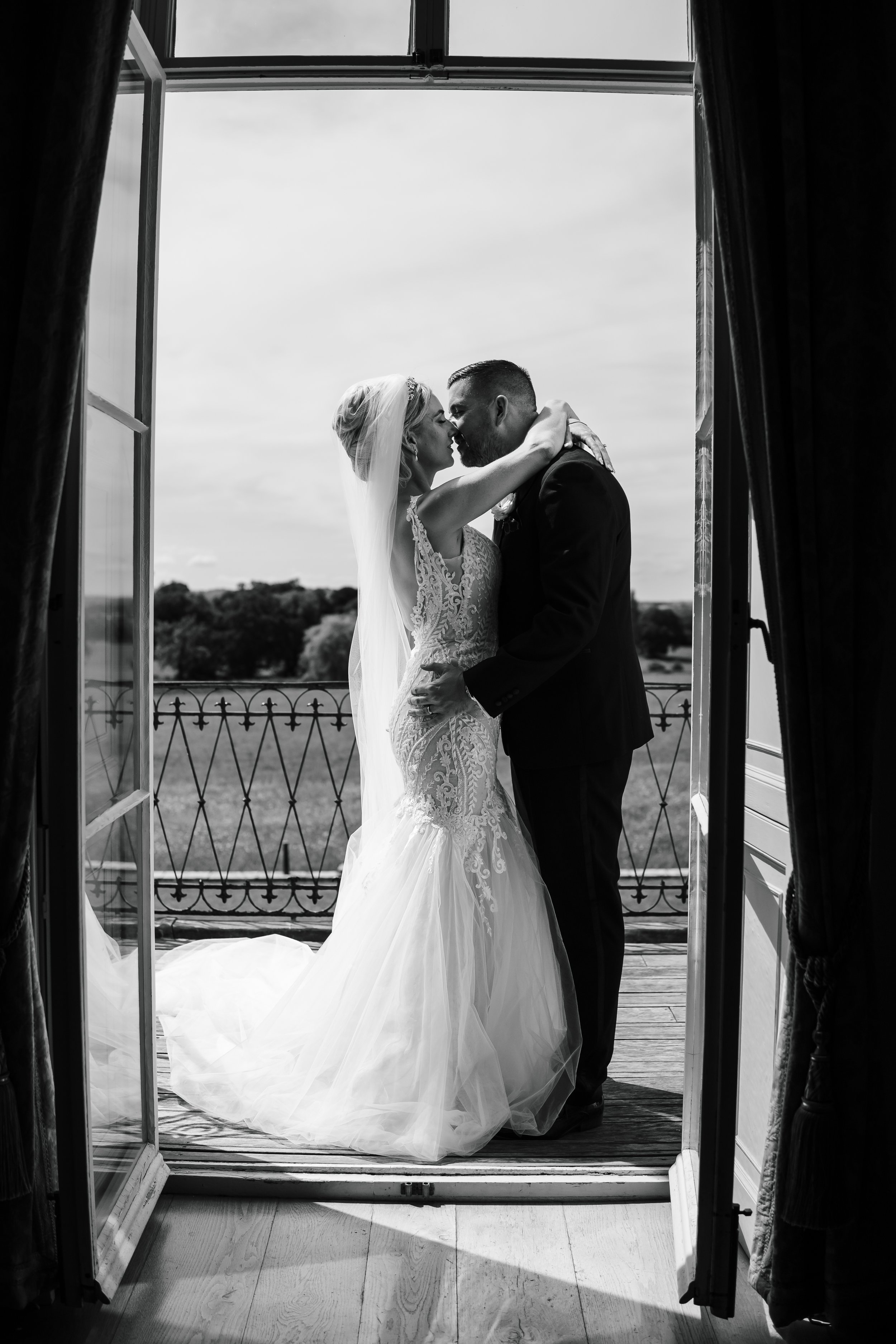 A bride and groom are sharing a kiss on a balcony, seen through open French doors. The bride is wearing a lace wedding gown and a veil, while the groom is in a dark suit. It's in black and white.