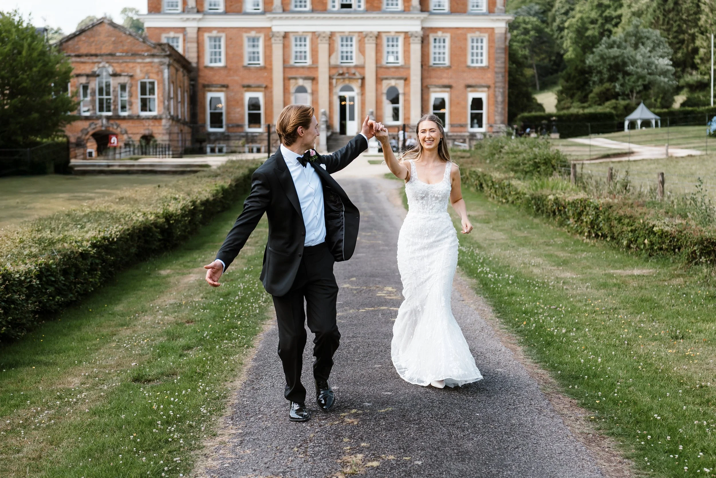 A newlywed couple, the groom in a black tuxedo and the bride in a white wedding gown, are dancing and smiling outdoors on a pathway in front of a large brick house with lush green surroundings.