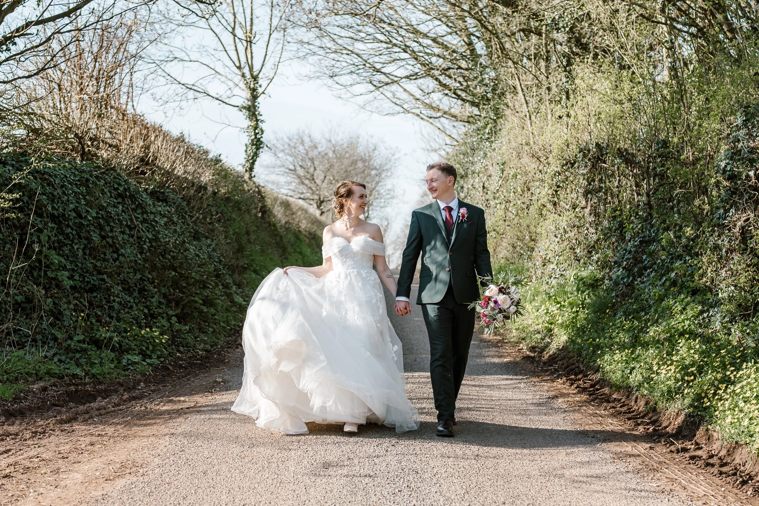A bride and groom walking hand in hand on a dirt pathway lined with trees and greenery, smiling at each other.