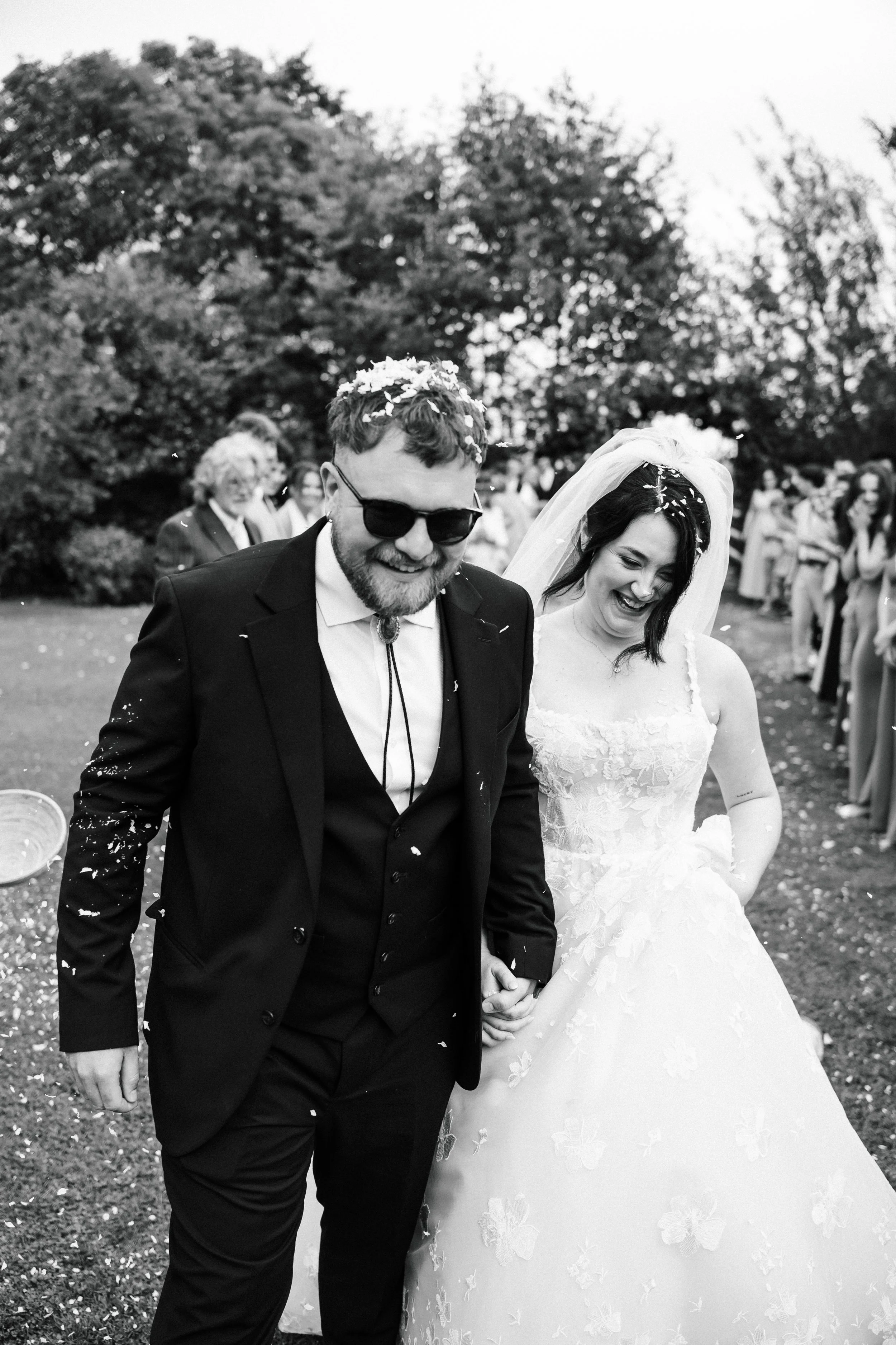 A happy couple walking outdoors on their wedding day, holding hands and smiling, surrounded by friends and family. The groom is wearing a black tuxedo and sunglasses, and the bride is wearing a white wedding dress and veil. Both are wearing flower cr