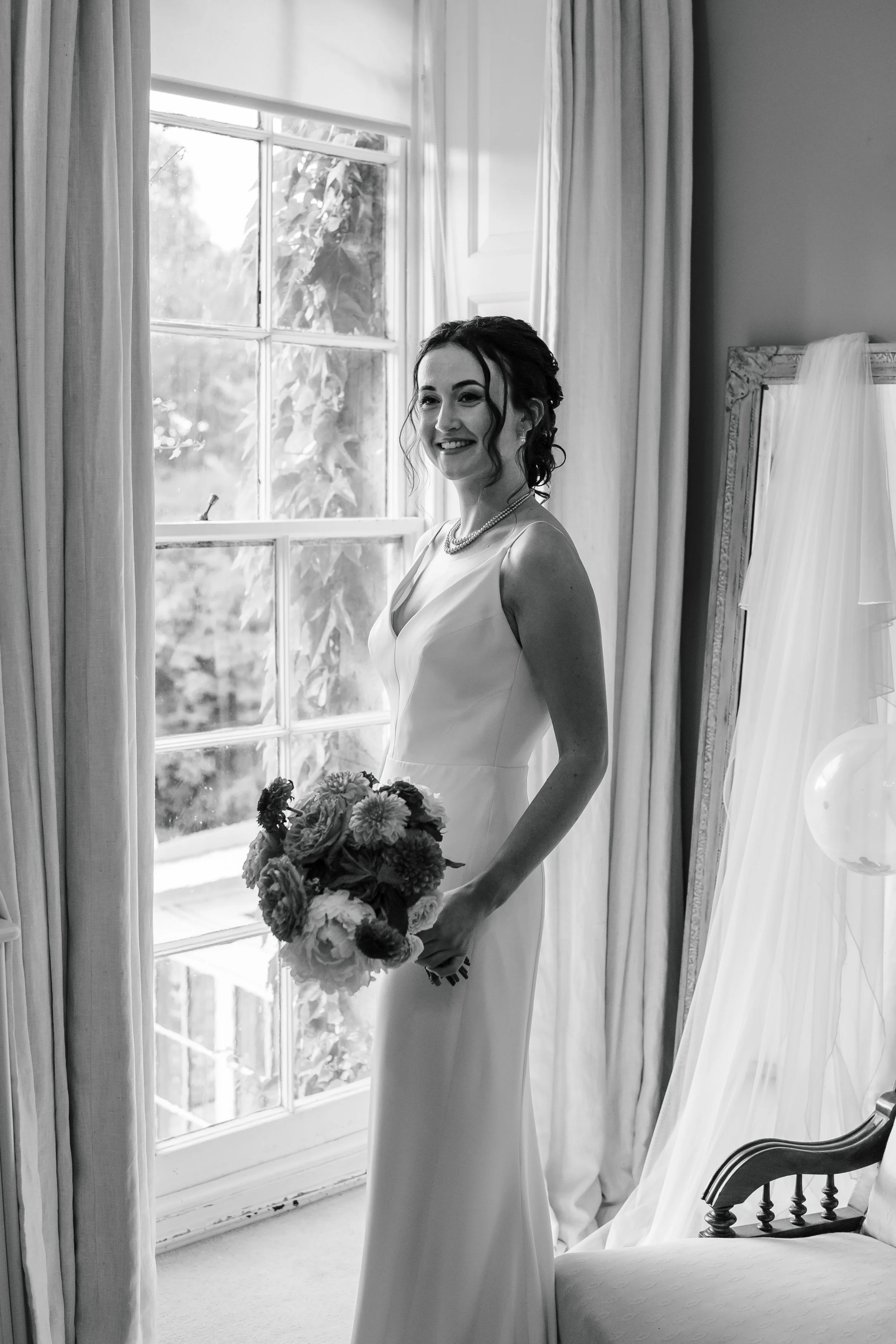 A smiling bride in a wedding dress holds a bouquet of flowers near a window with curtains and a mirror in the background.