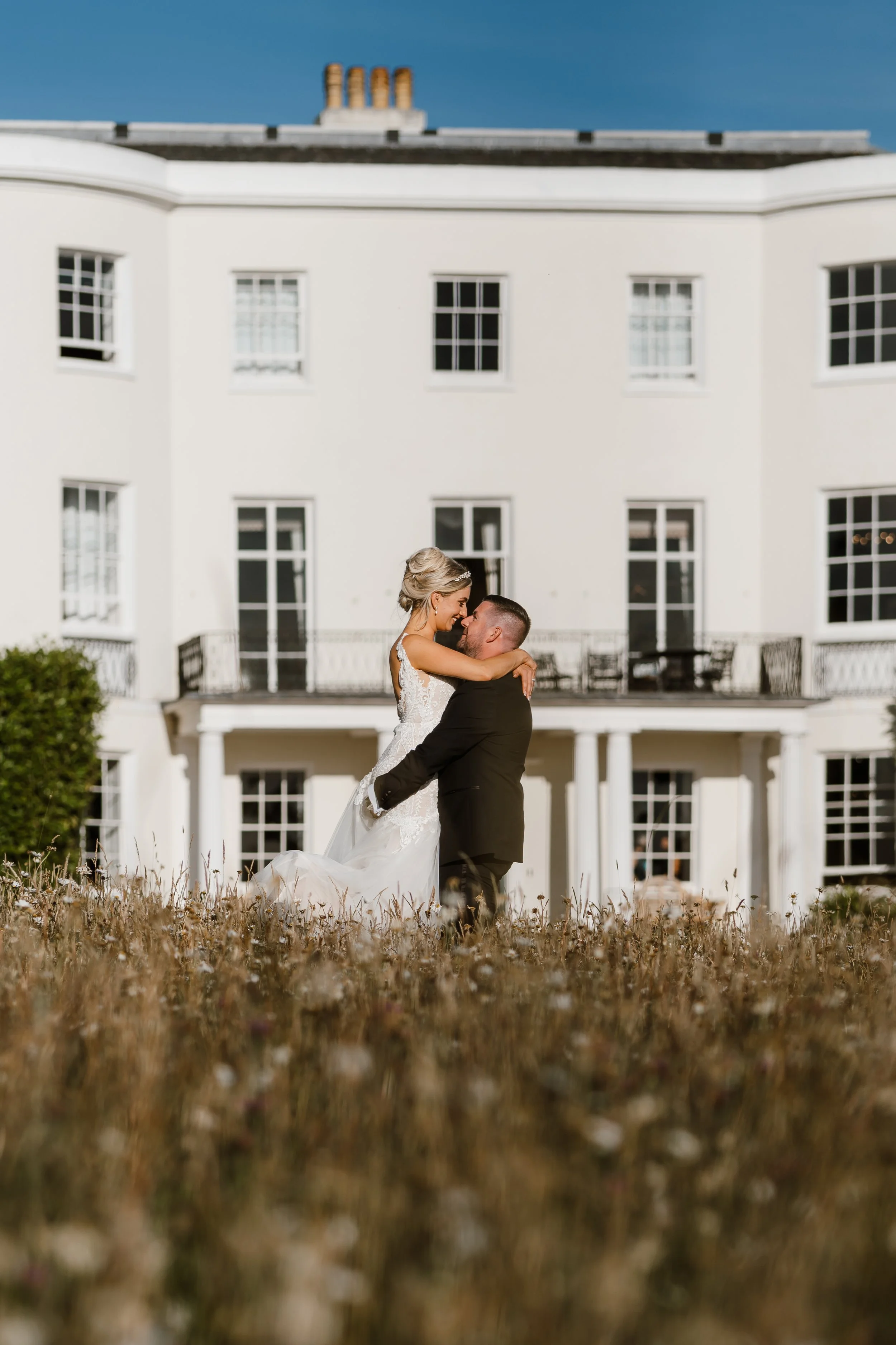 A bride and groom embracing in front of a white house with multiple windows and a balcony, outdoors on a sunny day.