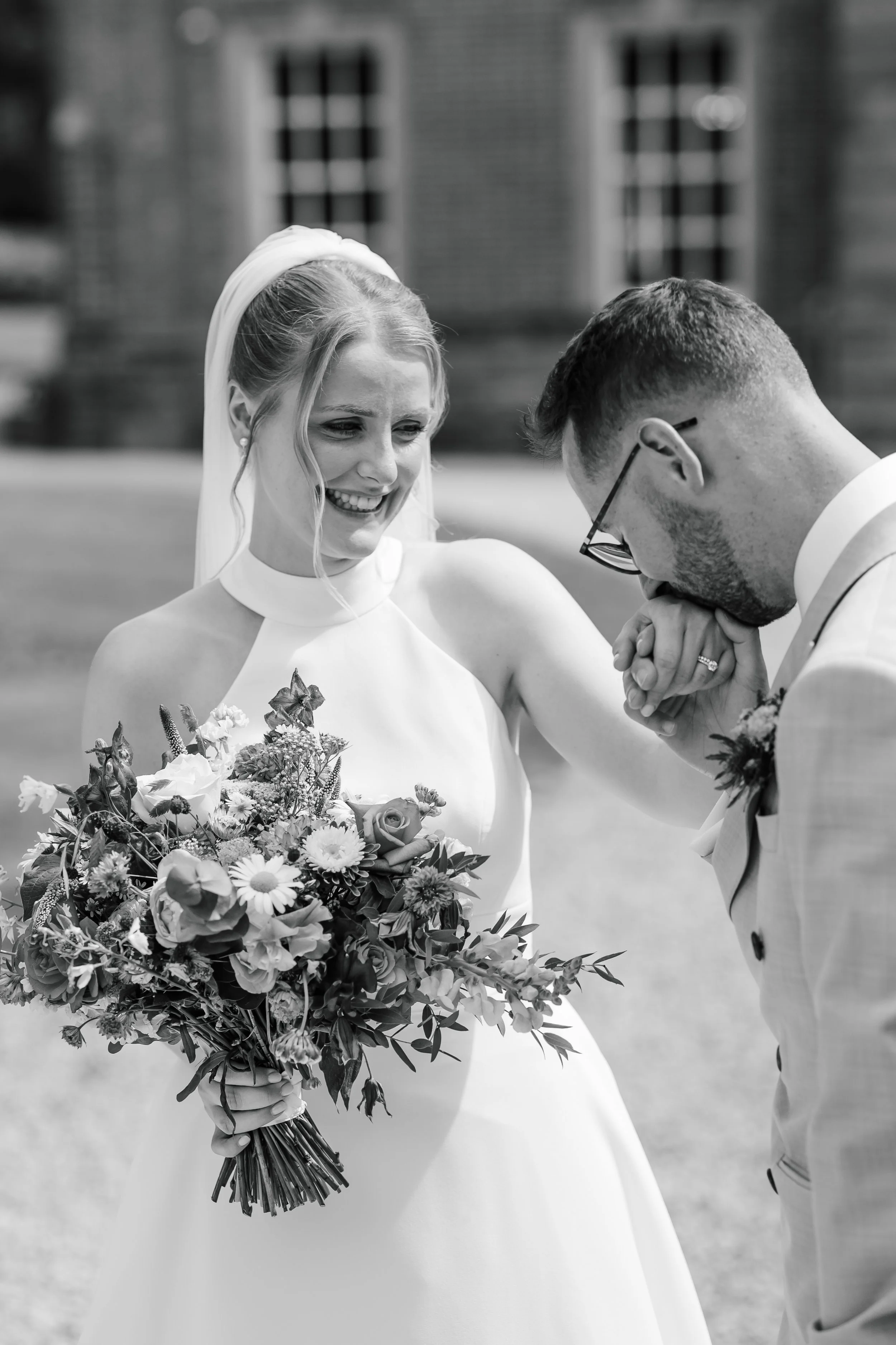 A bride in a white dress holding a large bouquet of flowers, smiling as a groom in glasses prays with her