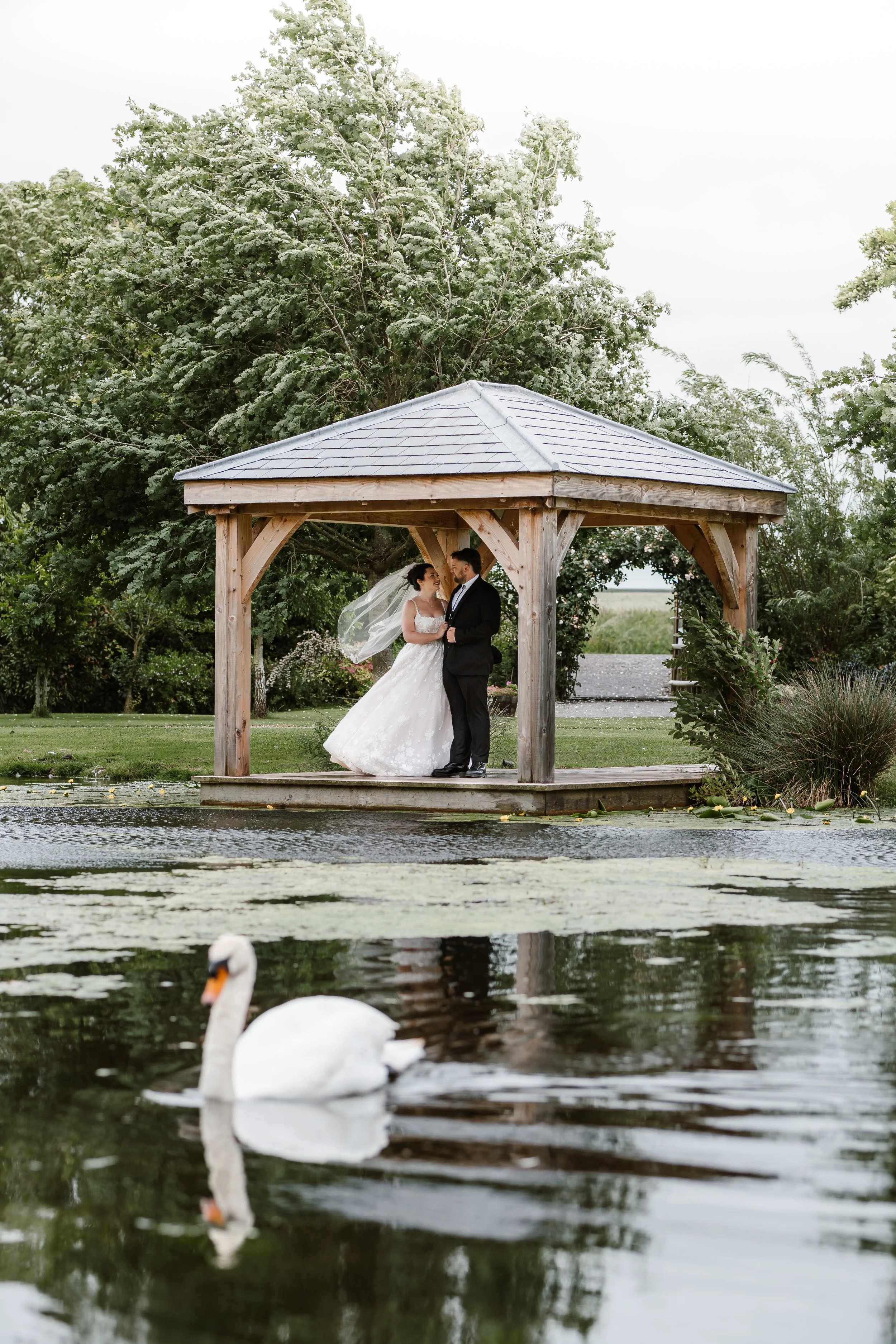 A bride and groom standing under a wooden gazebo surrounded by greenery, near a body of water with two swans swimming nearby.