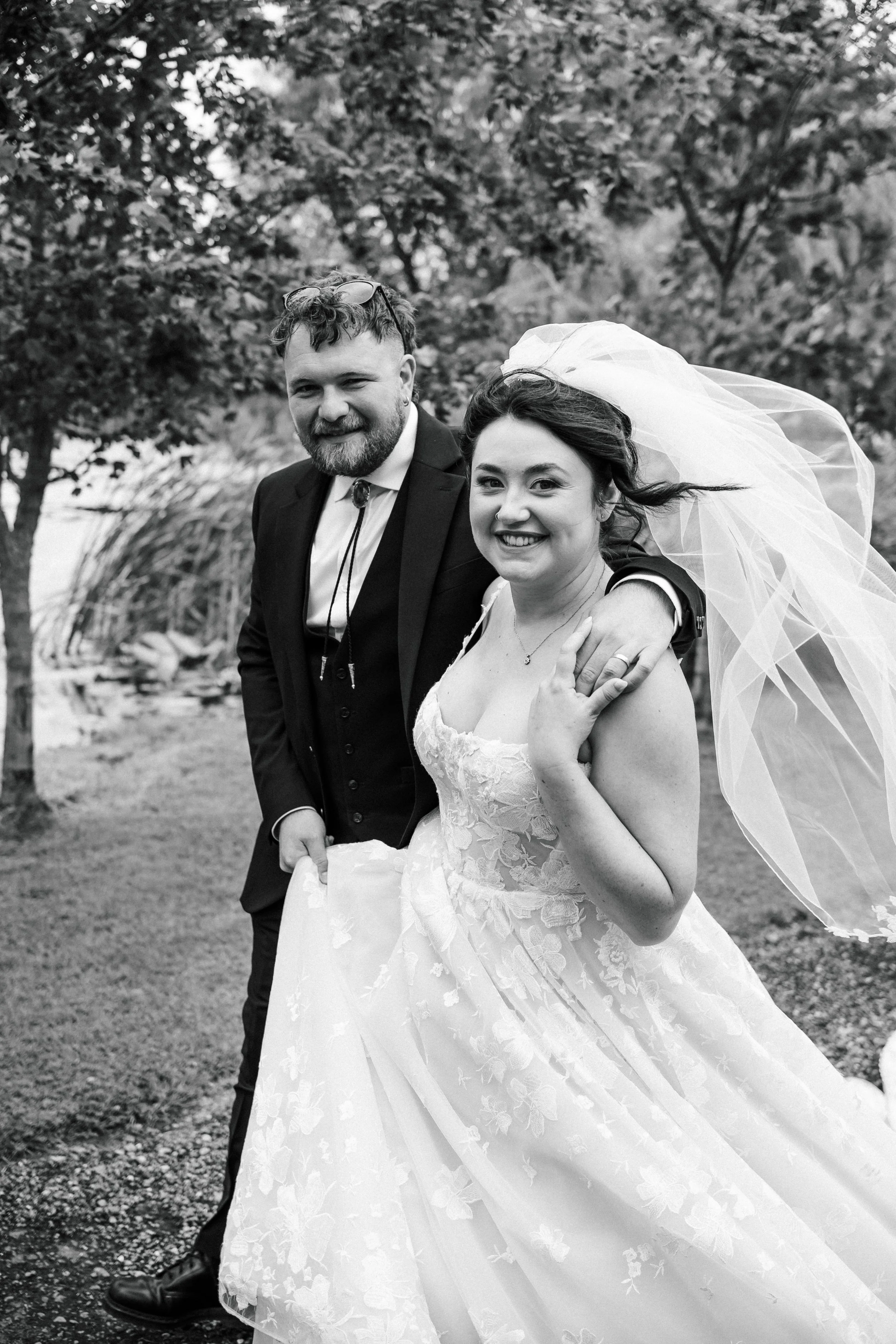 Black and white photo of a newlywed couple outdoors, smiling, with trees and grass in the background. The woman is wearing a lace wedding dress and veil, and the man is in a suit with a bolo tie.