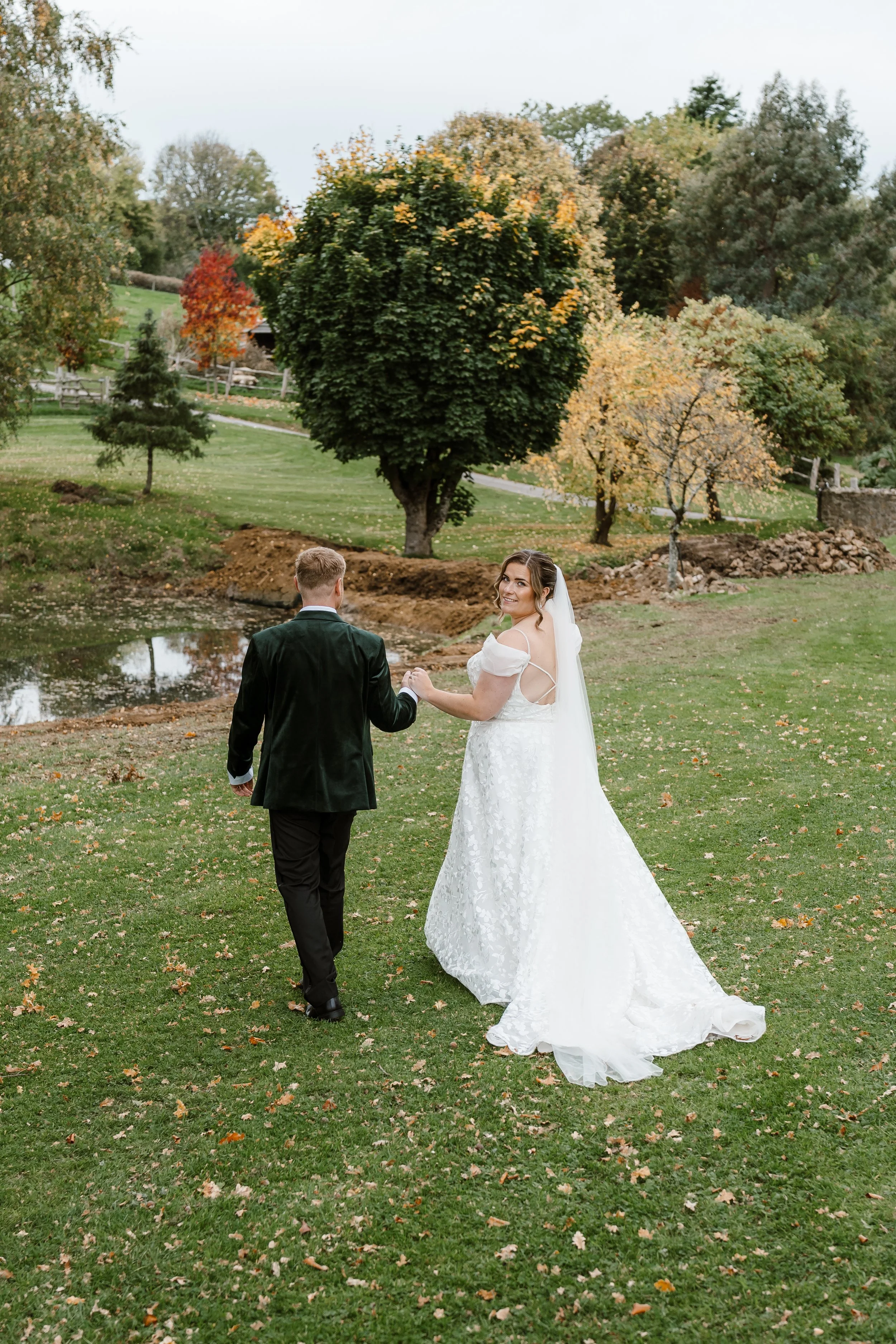 Bride and groom holding hands and walking outdoors on their wedding day, surrounded by trees with fall foliage, near a small pond.