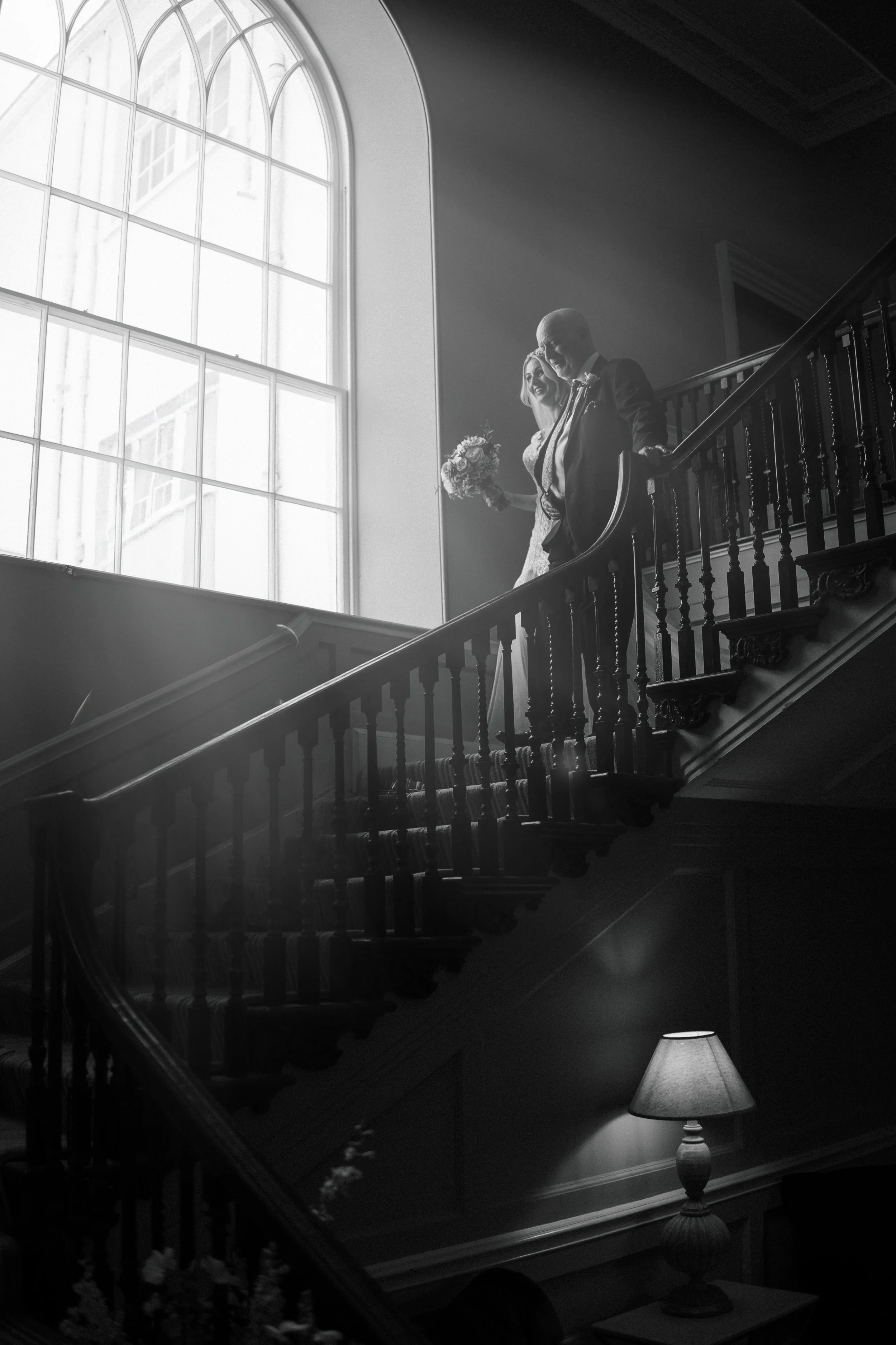 A bride and groom standing on a staircase inside a building with large arched windows, holding bouquets, dressed in wedding attire, with light streaming through the windows.
