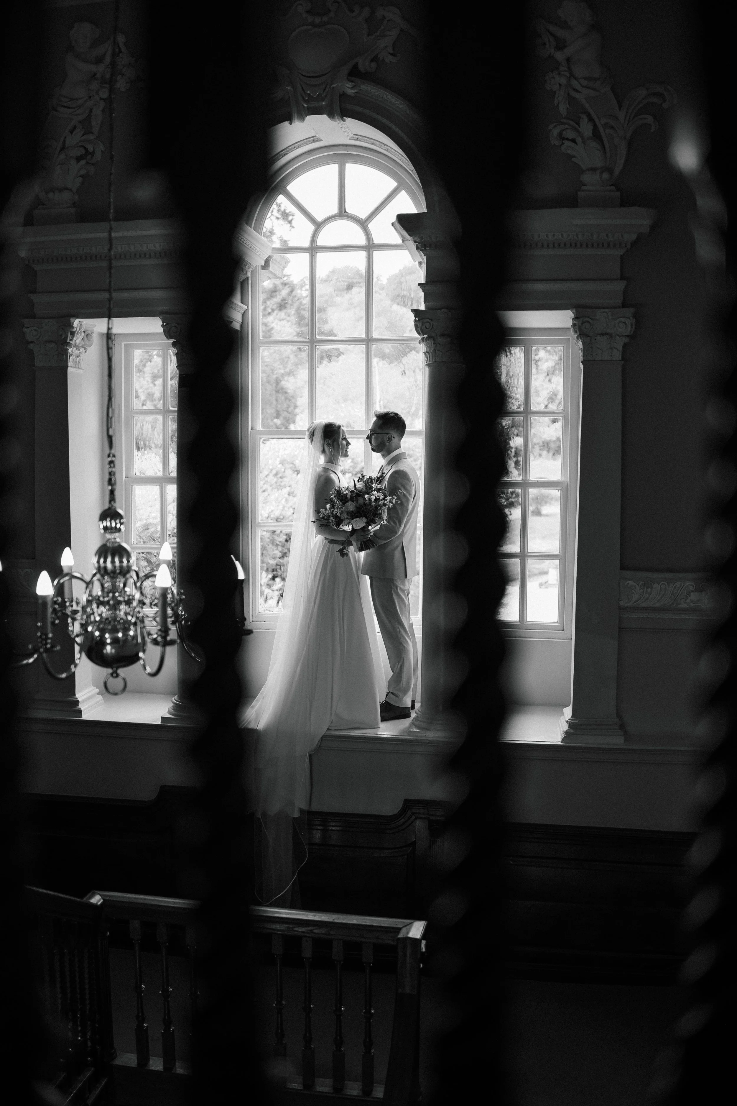 A bride and groom standing face to face in front of a large arched window inside a grand, ornate building, possibly a church or mansion, with decorative columns. They are holding a bouquet together, and the scene is captured through wrought iron bars