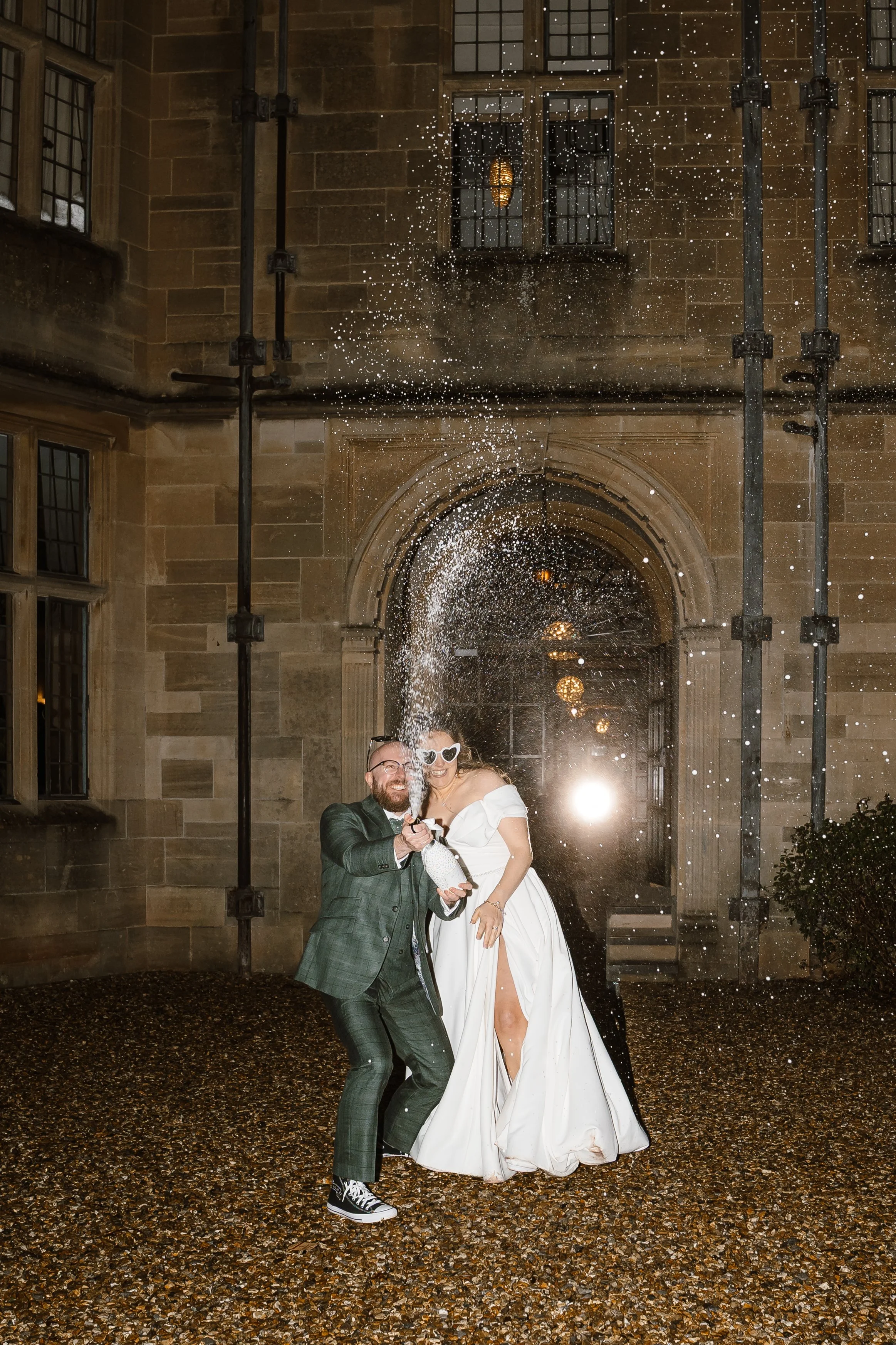 A couple celebrating at their wedding, standing outside a stone building, spraying champagne and smiling.