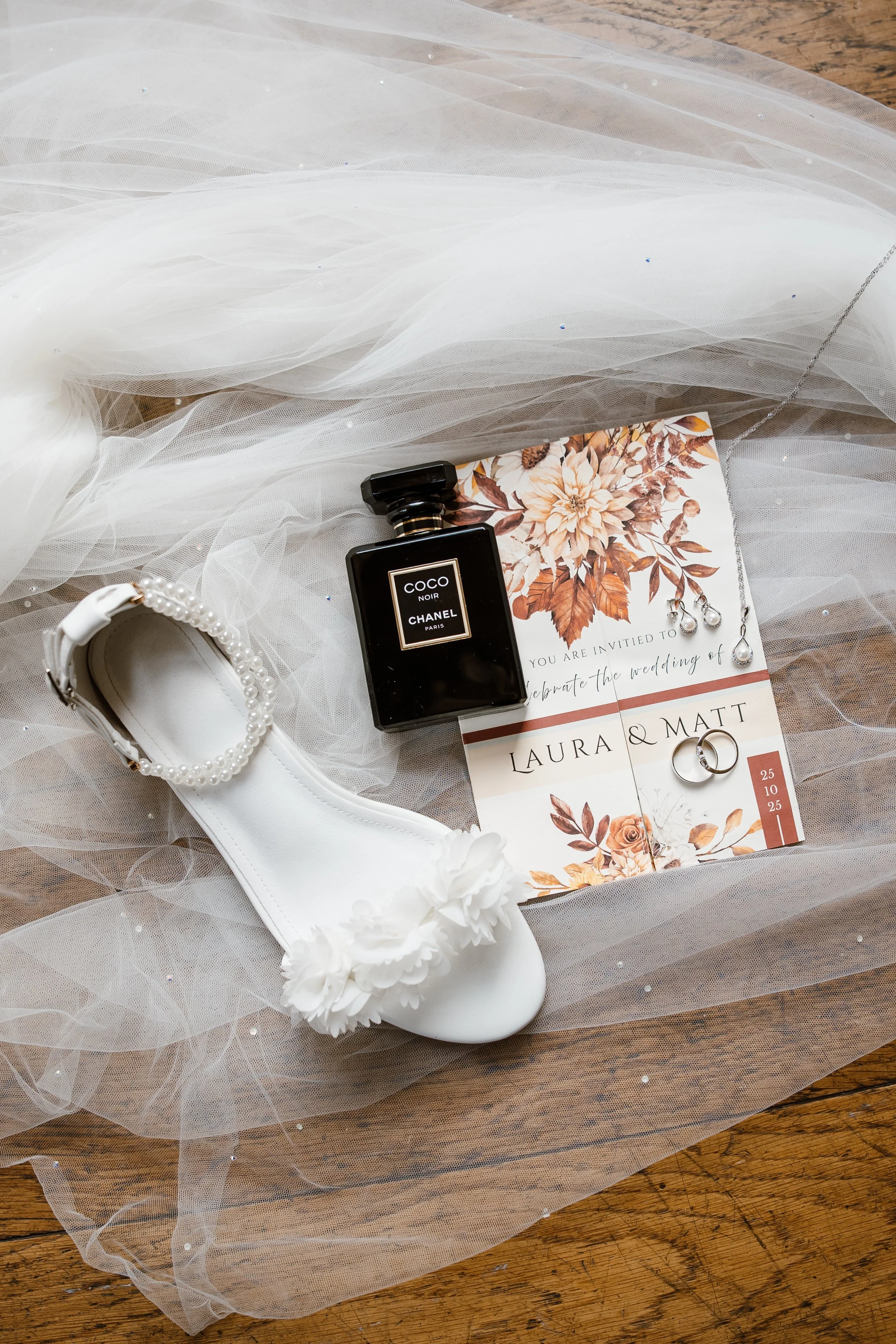Wedding invitation surrounded by wedding accessories including a white lace shoe decorated with pearls and flowers, a bottle of Chanel perfume, a pearl bracelet, and a pair of wedding rings, all placed on a sheer tulle fabric on a wooden surface.