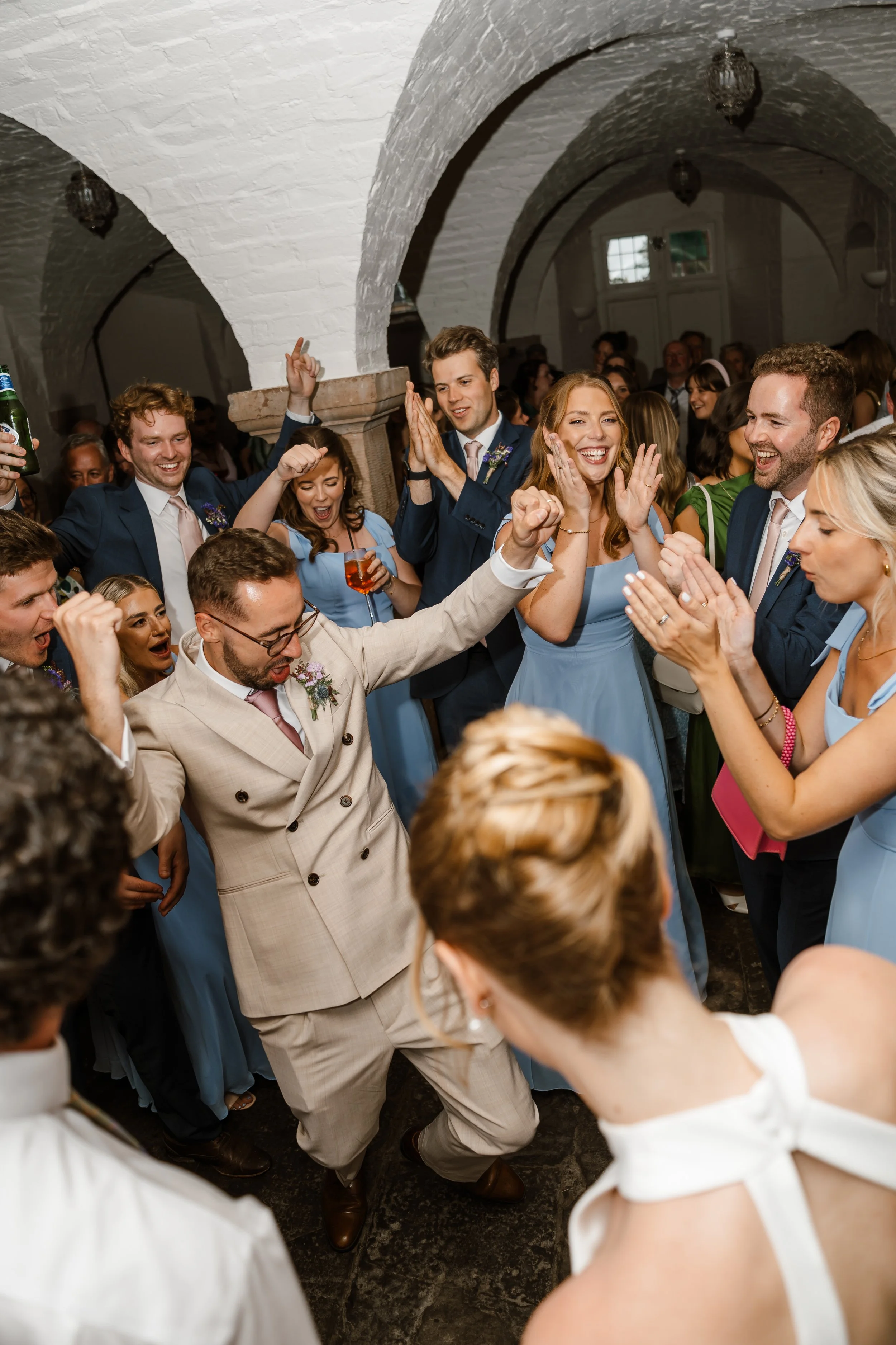 People at a wedding reception dancing and celebrating in a historic stone building with arches, white brick walls, and lantern-style lights.