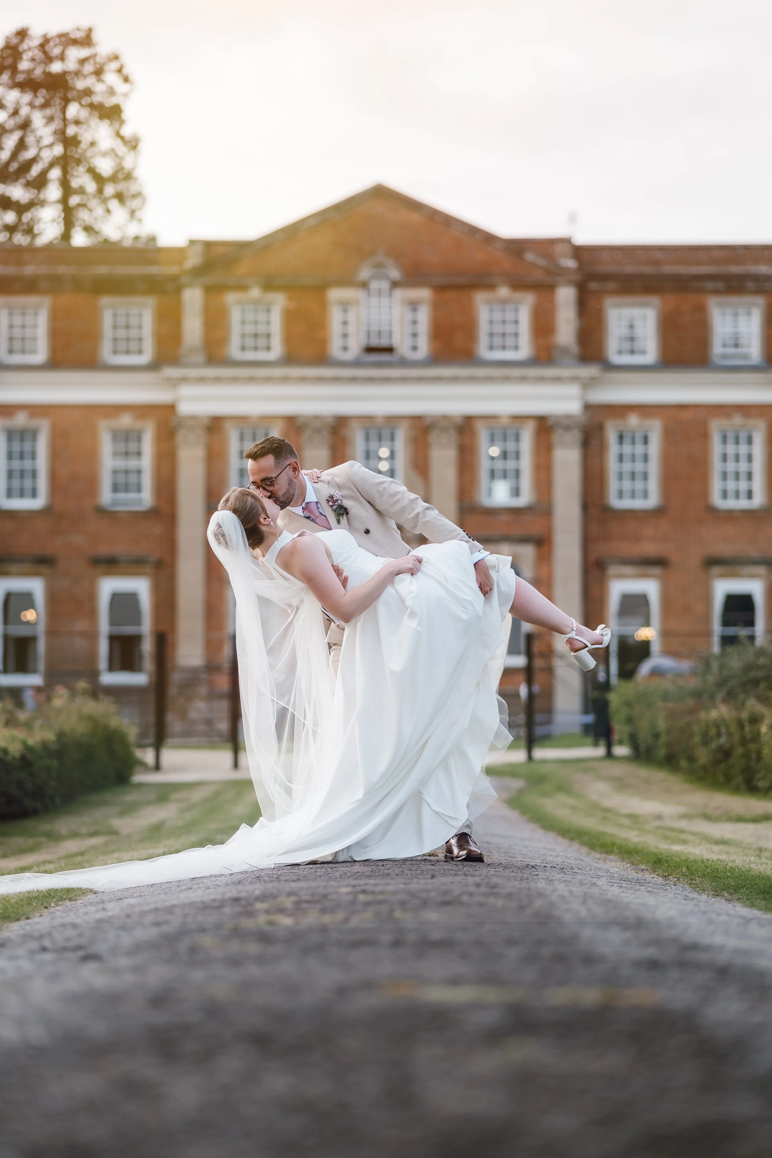 A newlywed couple sharing a kiss outside a large brick mansion, with the groom dipping the bride in a romantic pose, as the sun sets behind them.
