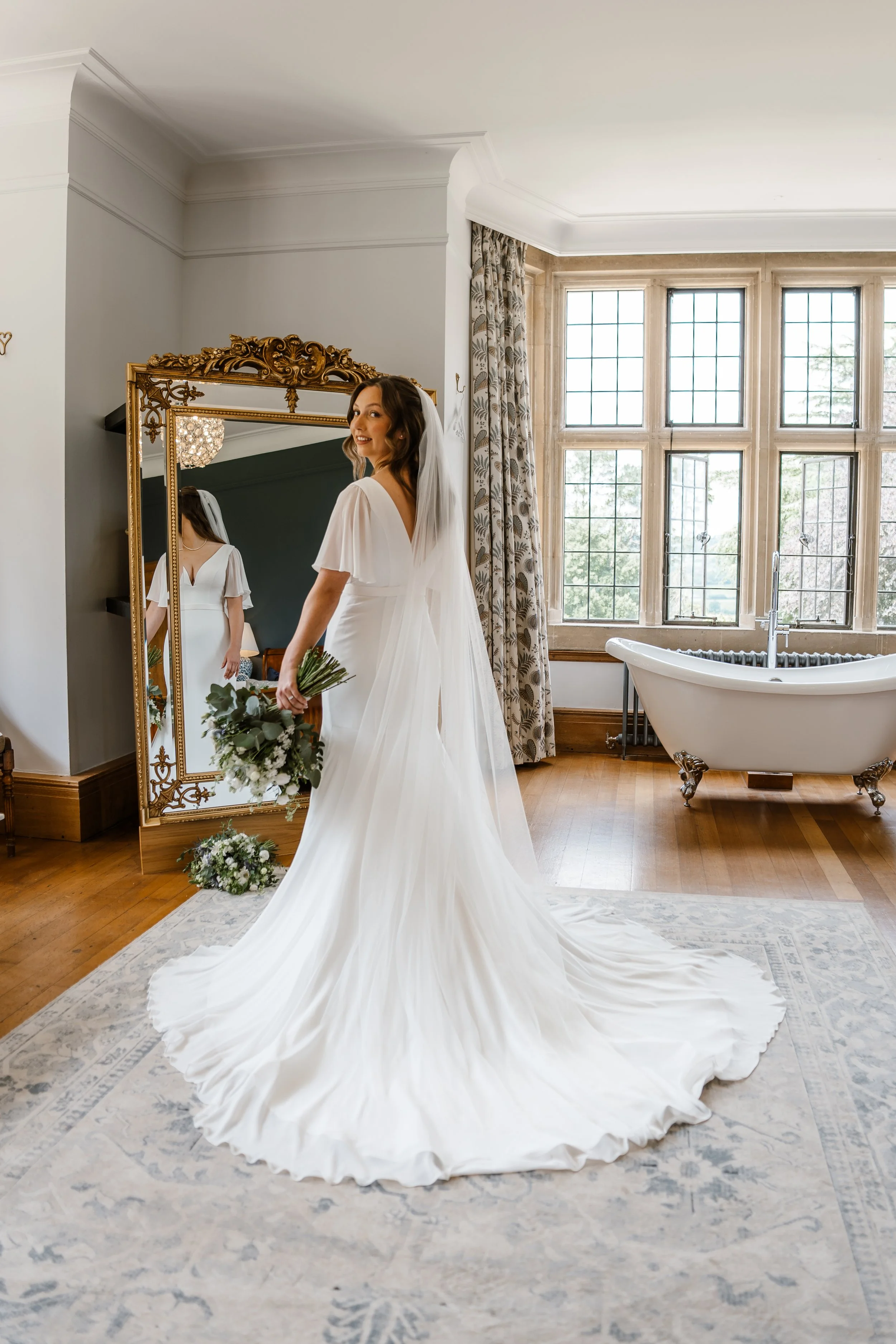 A bride in a flowing white wedding gown and veil stands in a bright room with large windows, holding a bouquet of flowers while looking into a gold-framed mirror.