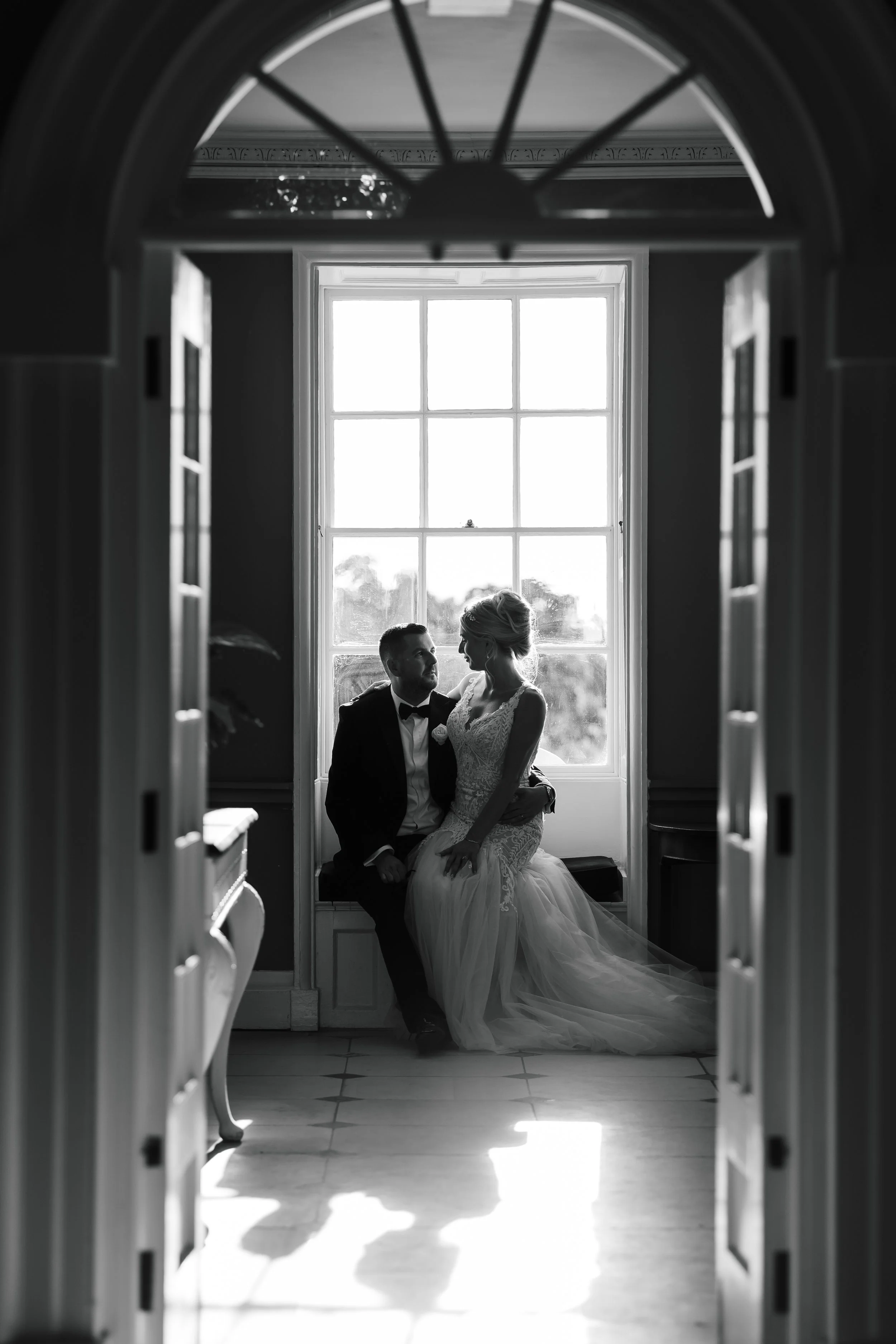 Black and white photo of a bride and groom sitting together by a large window, viewed through an archway with shelves on either side. The bride is in a lace wedding dress, and the groom is in a tuxedo with a bow tie. They are looking at each other, w
