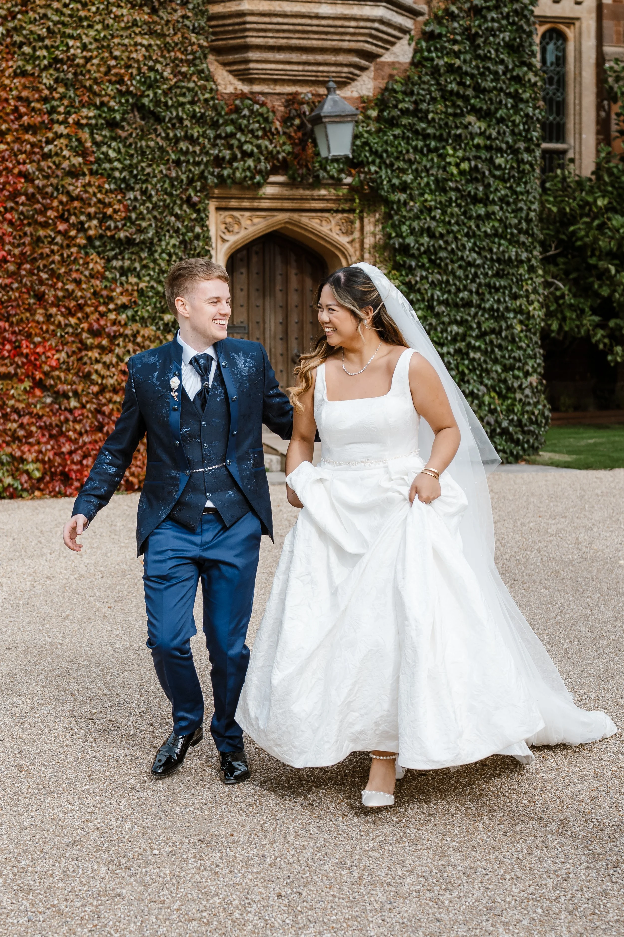 A newlywed couple, a man and woman, smiling and walking together outside in front of a historic building covered in ivy, during their wedding day.