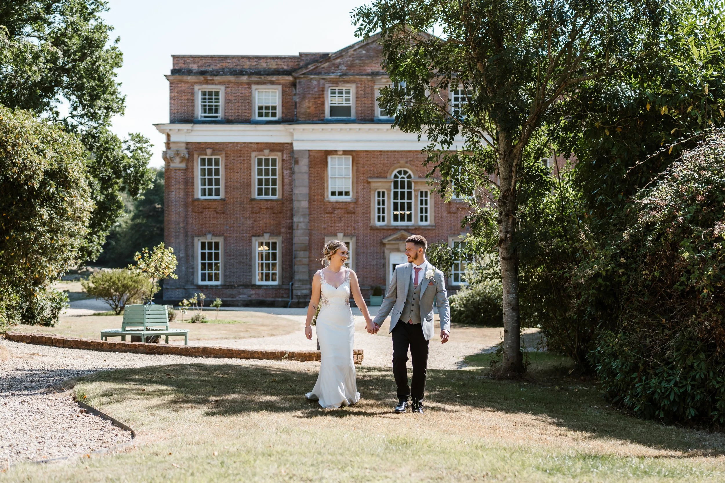 A bride and groom holding hands while walking outside on a sunny day near a large brick building surrounded by trees and greenery.