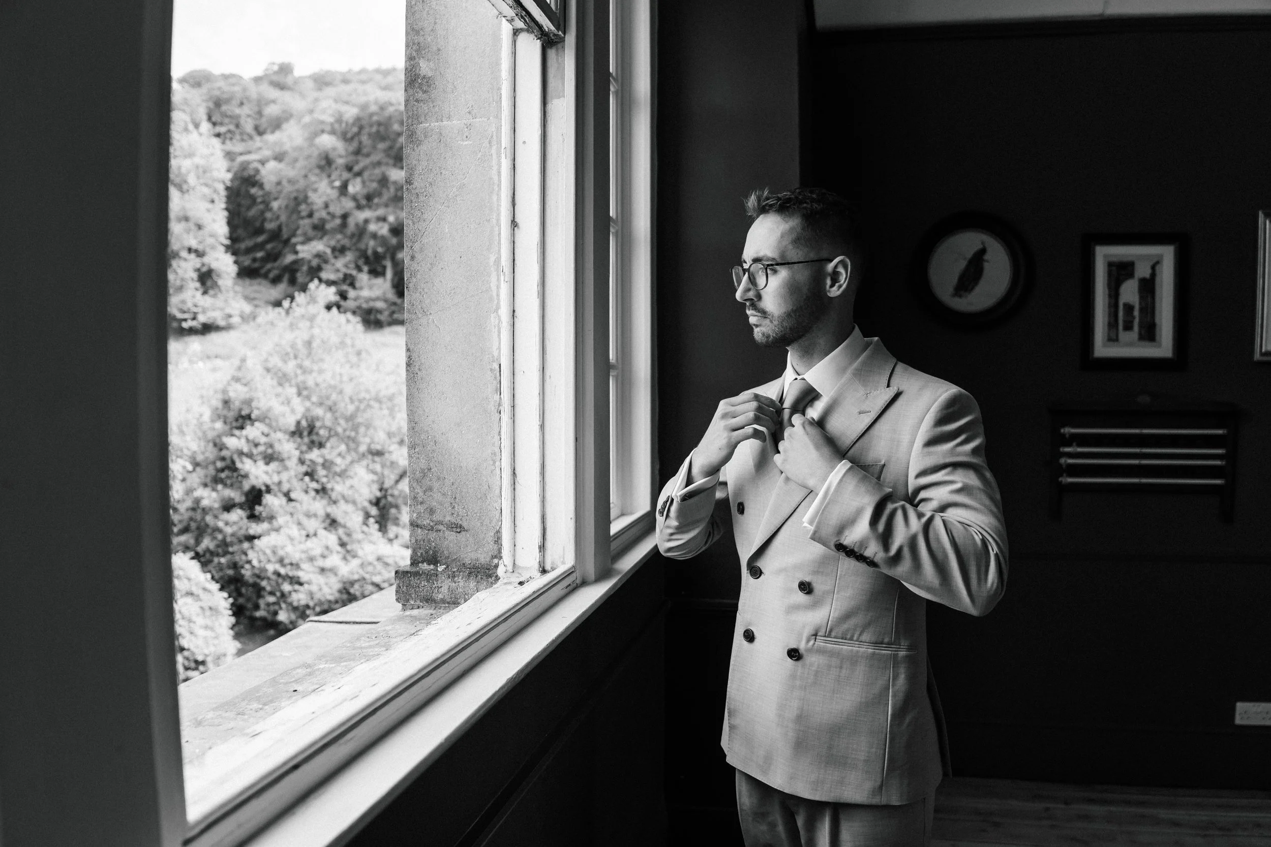 A man in a light-colored suit adjusting his tie while standing by a window, with outdoor greenery visible outside.
