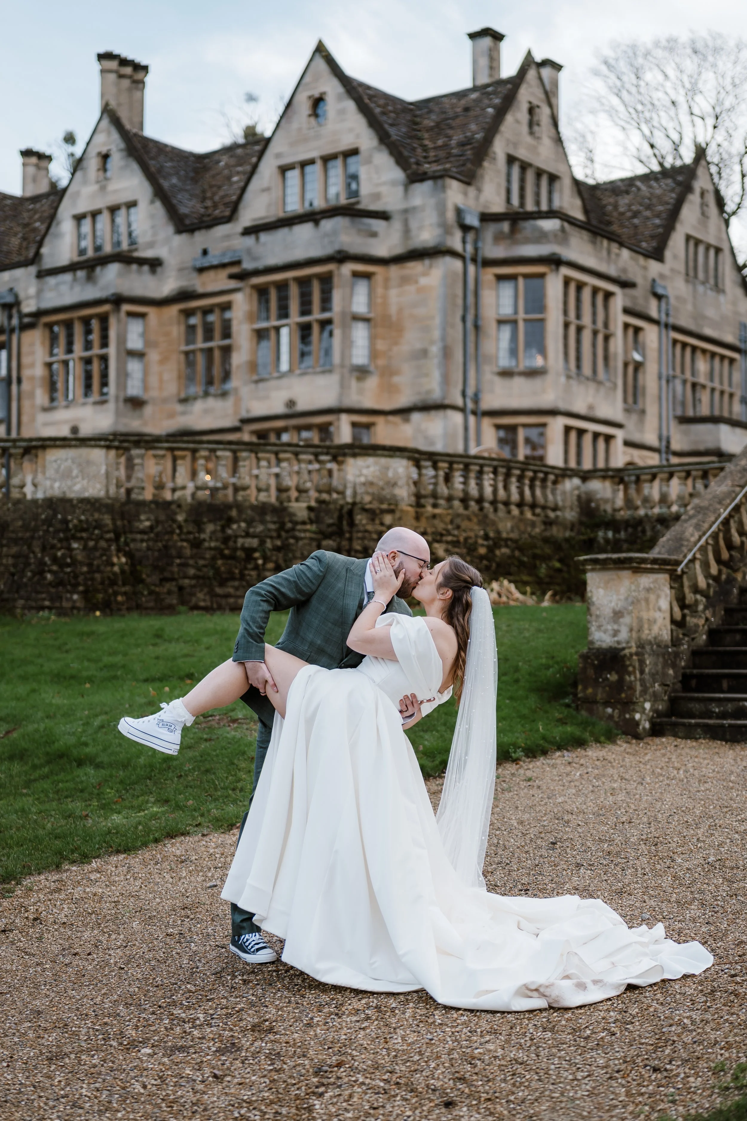 A bride and groom kissing outdoors, with the groom holding the bride in his arms, in front of a historic stone building with multiple gables and windows.