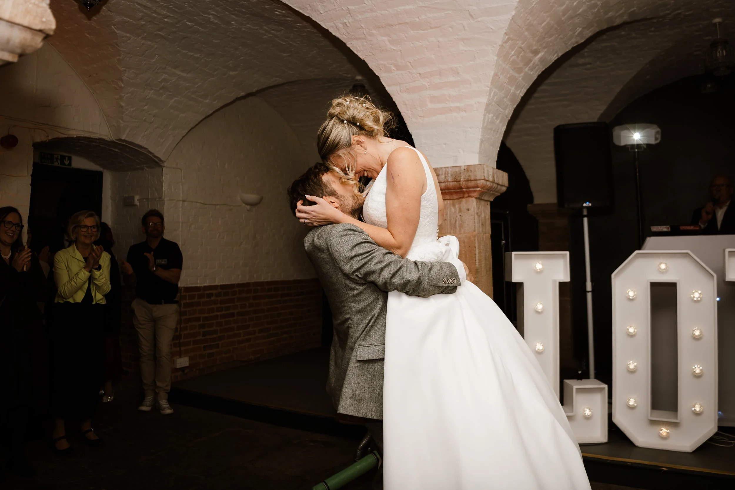 A bride and groom are sharing a dance at their wedding reception. The groom is lifting the bride, who is leaning towards him, in a joyful moment. Some guests are clapping and smiling in the background, inside a brick-walled venue with arched ceilings
