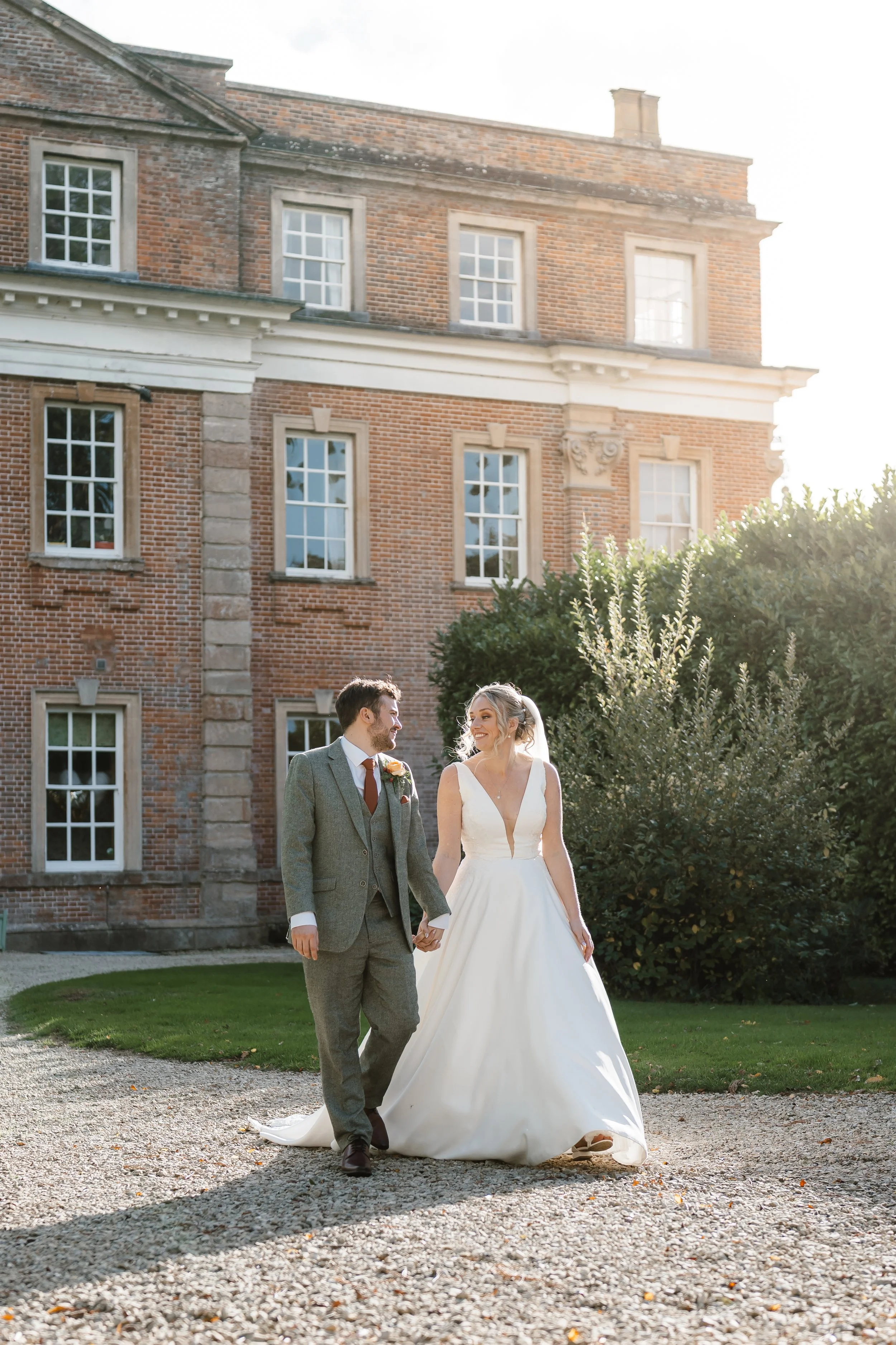 A bride and groom walking hand in hand on a gravel path outside a brick building during sunset.