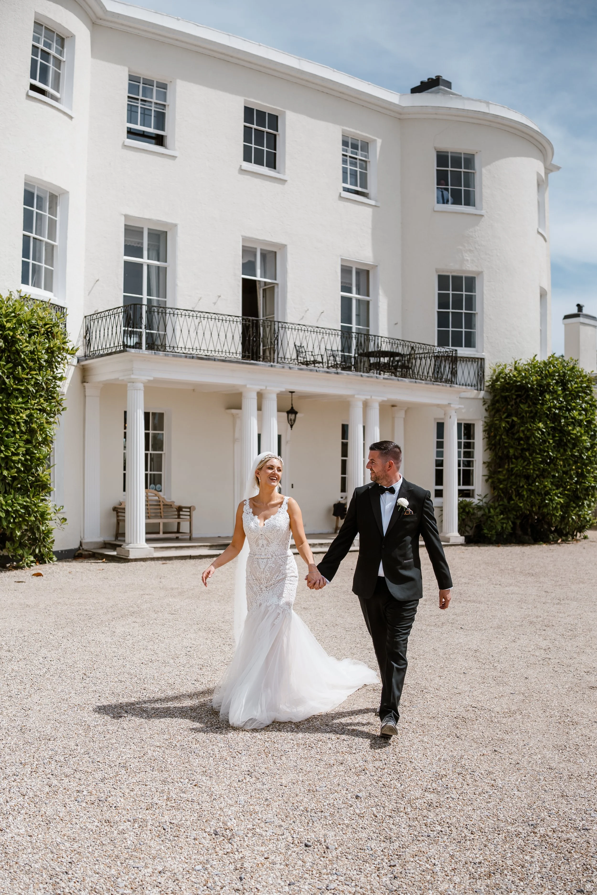 A bride and groom holding hands and walking outside a white mansion on a sunny day.