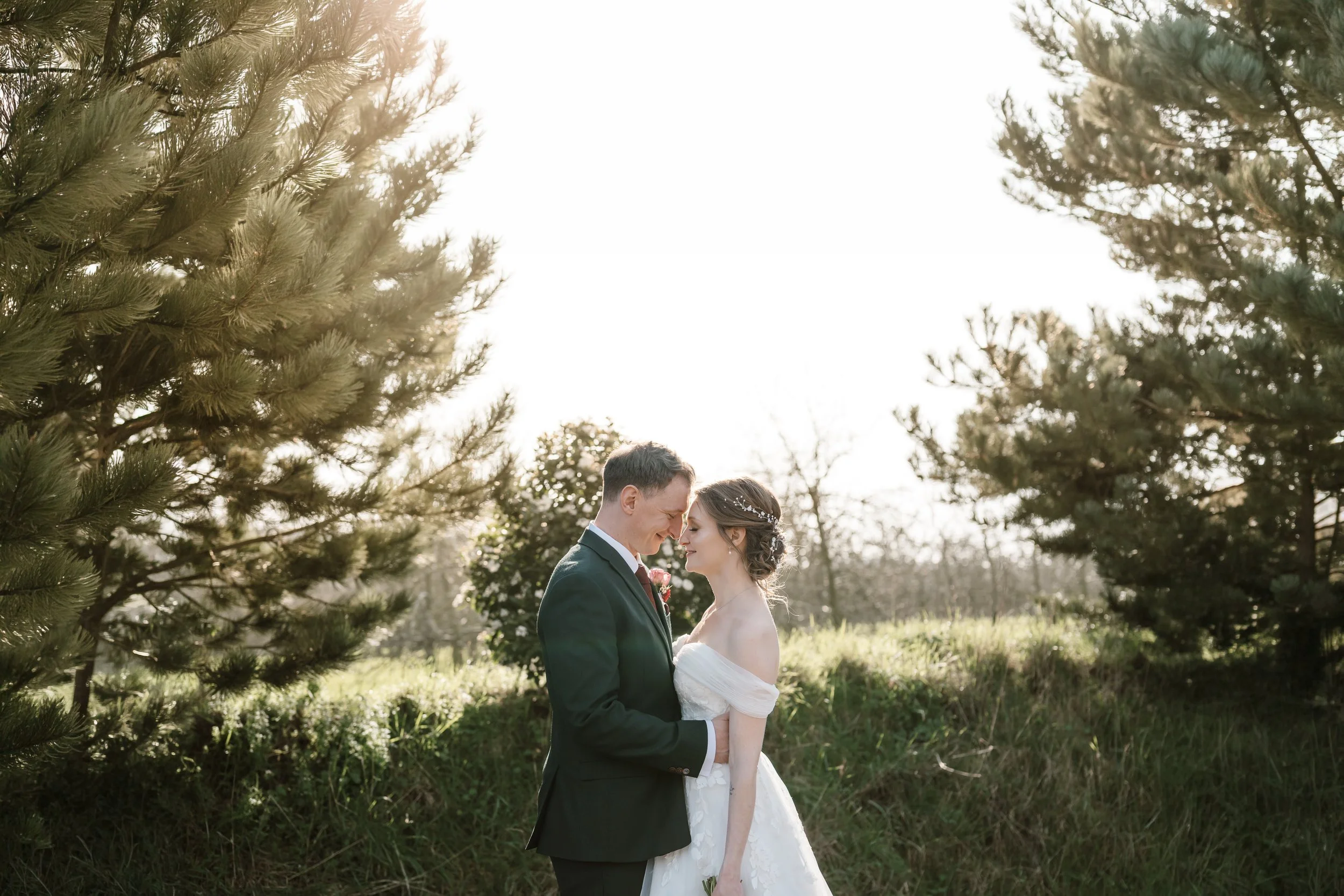 A bride and groom stand close together outdoors, touching foreheads and smiling, surrounded by trees and greenery, during what appears to be a wedding photoshoot.