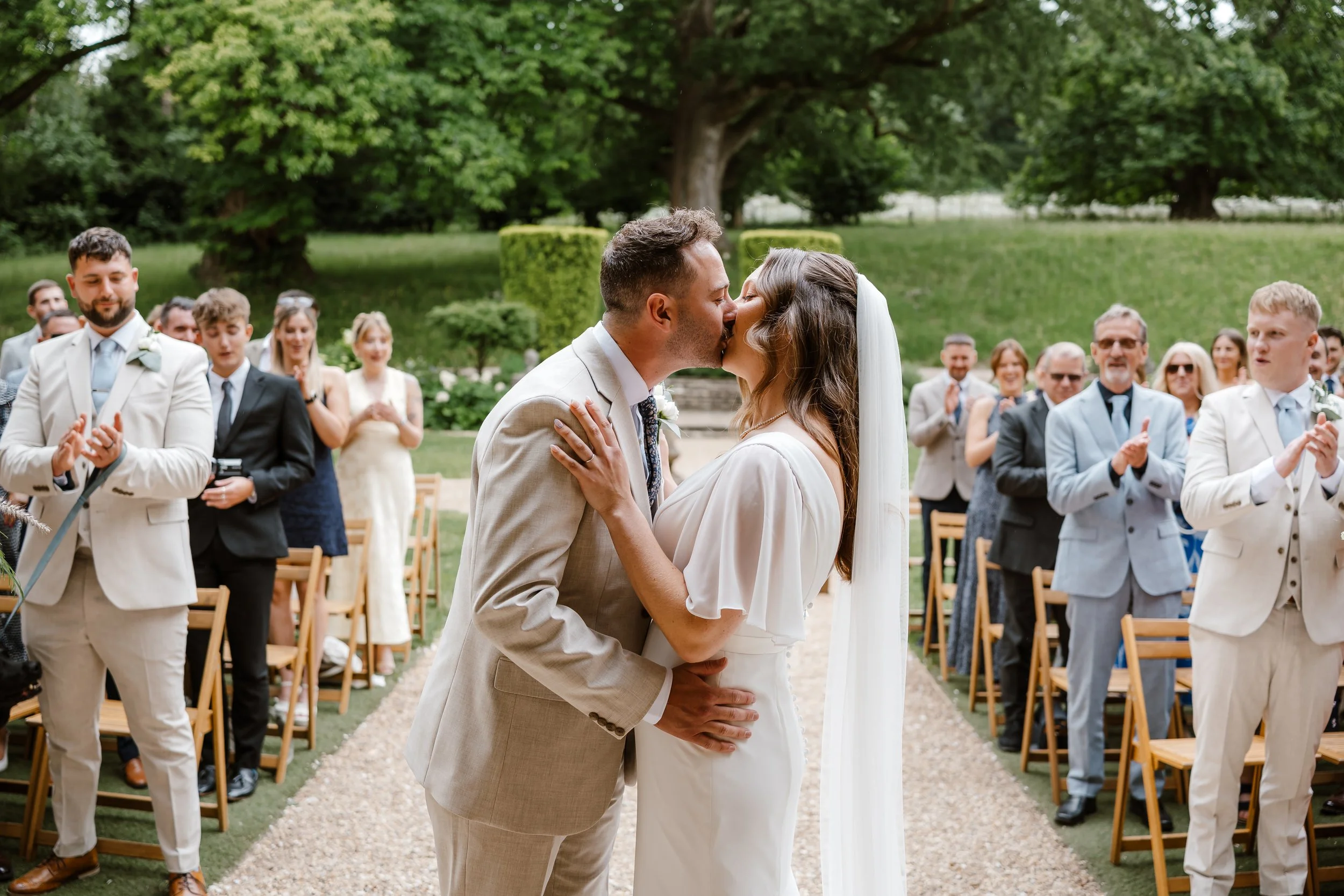 A wedding ceremony outdoors with a bride and groom kissing, surrounded by friends and family clapping and celebrating, with green trees in the background.