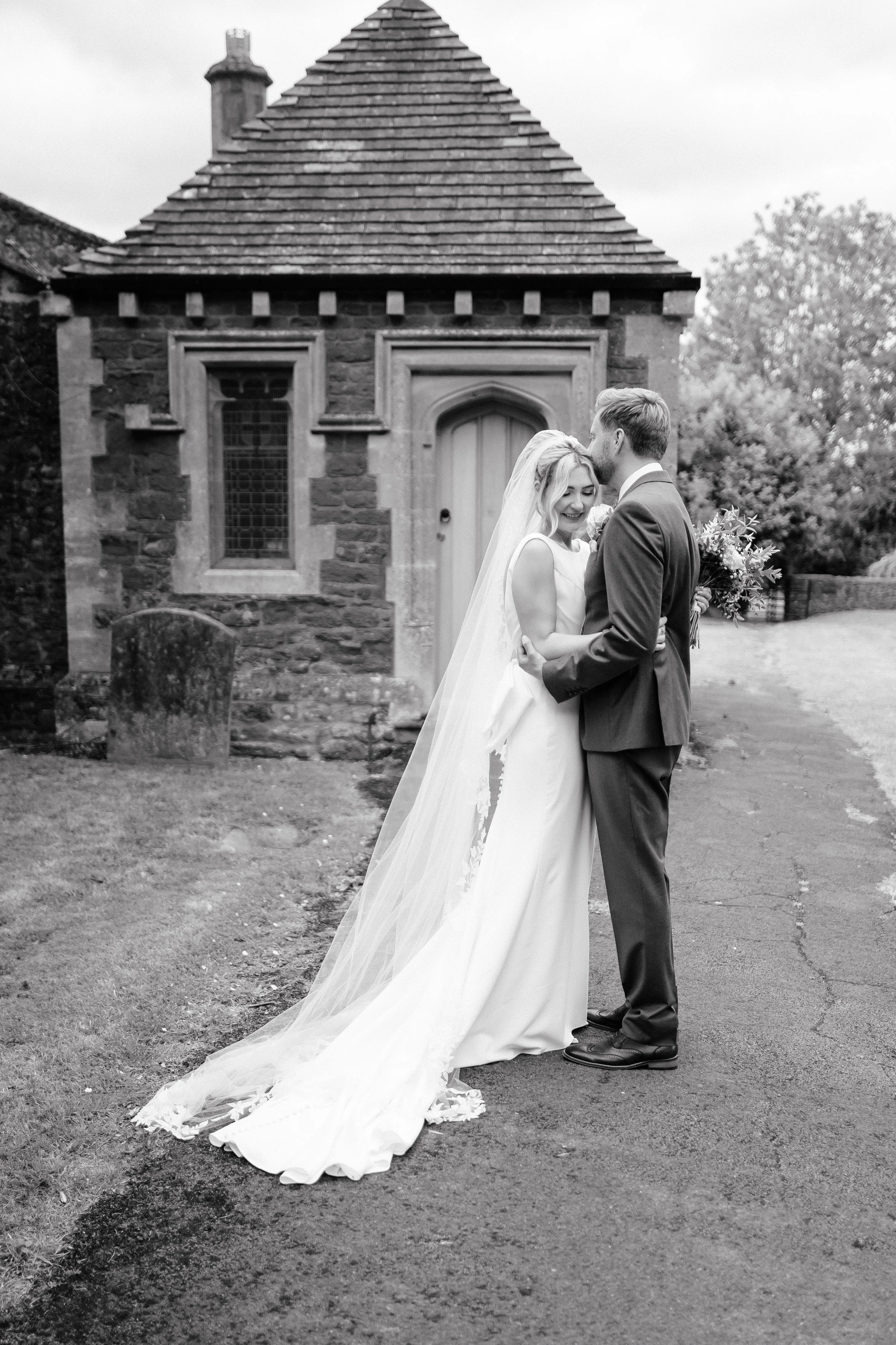 Black and white photo of a bride and groom embracing outdoors, with a small stone building featuring a pitched roof in the background.