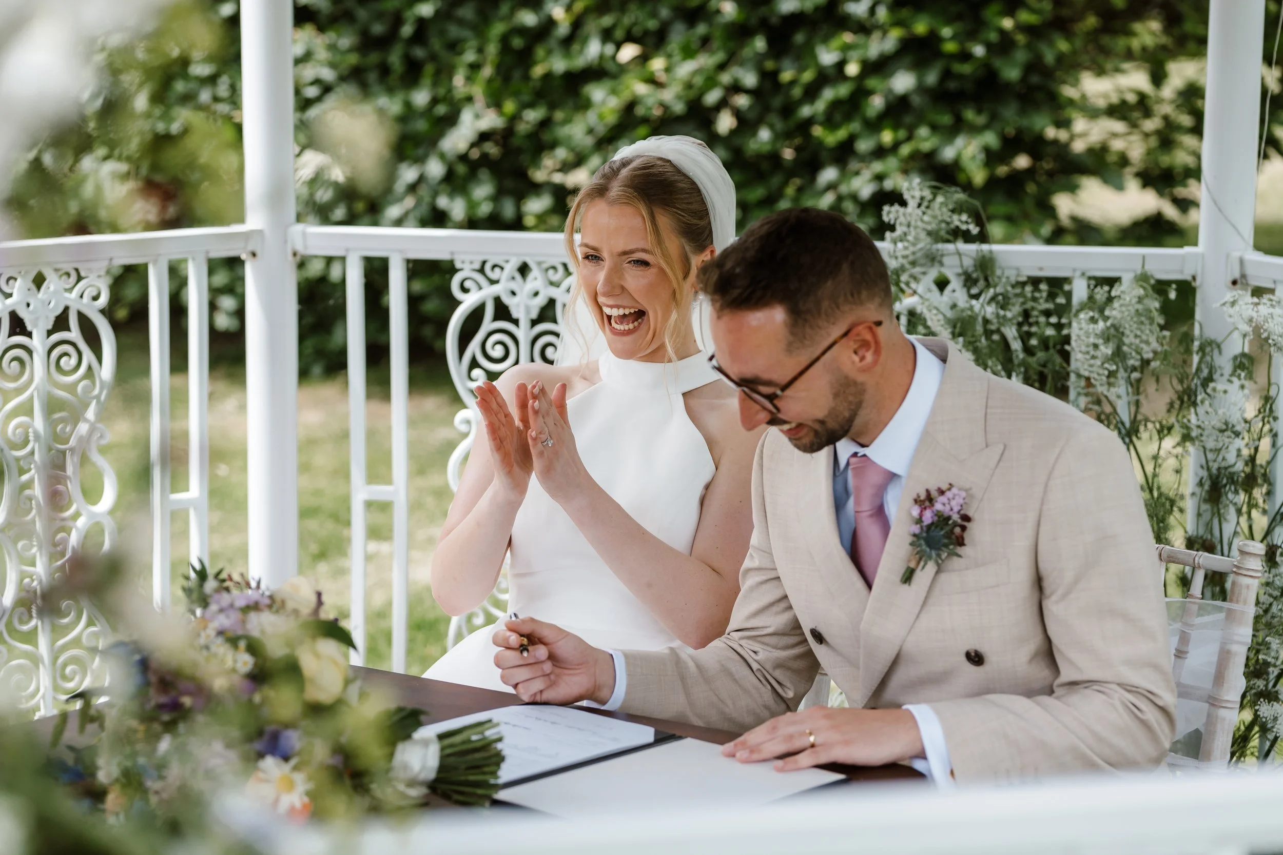 A bride and groom, seated at a table outdoors, are sharing a joyful moment during their wedding ceremony. The bride is smiling and clapping, while the groom is looking down, both dressed in wedding attire.