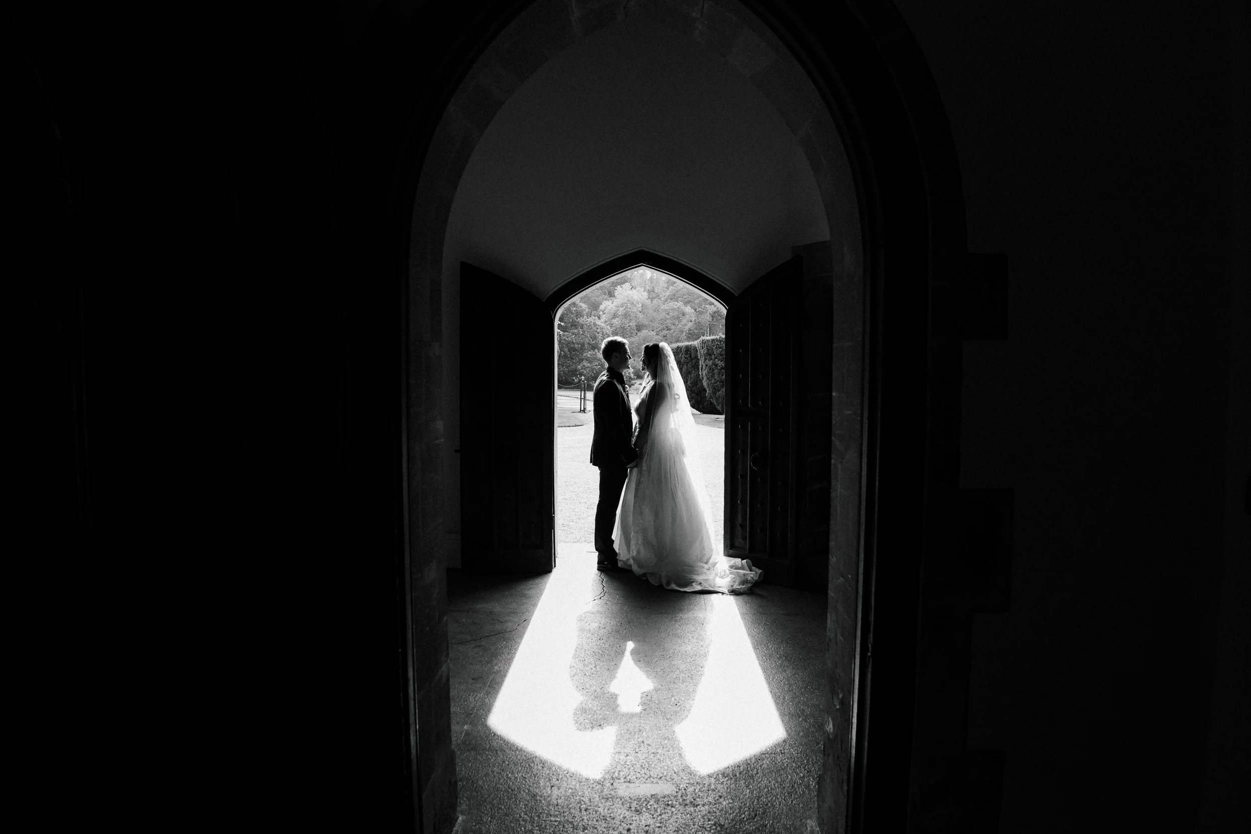 Black and white photo of a bride and groom holding hands and facing each other in a doorway, with outdoor scenery in the background.