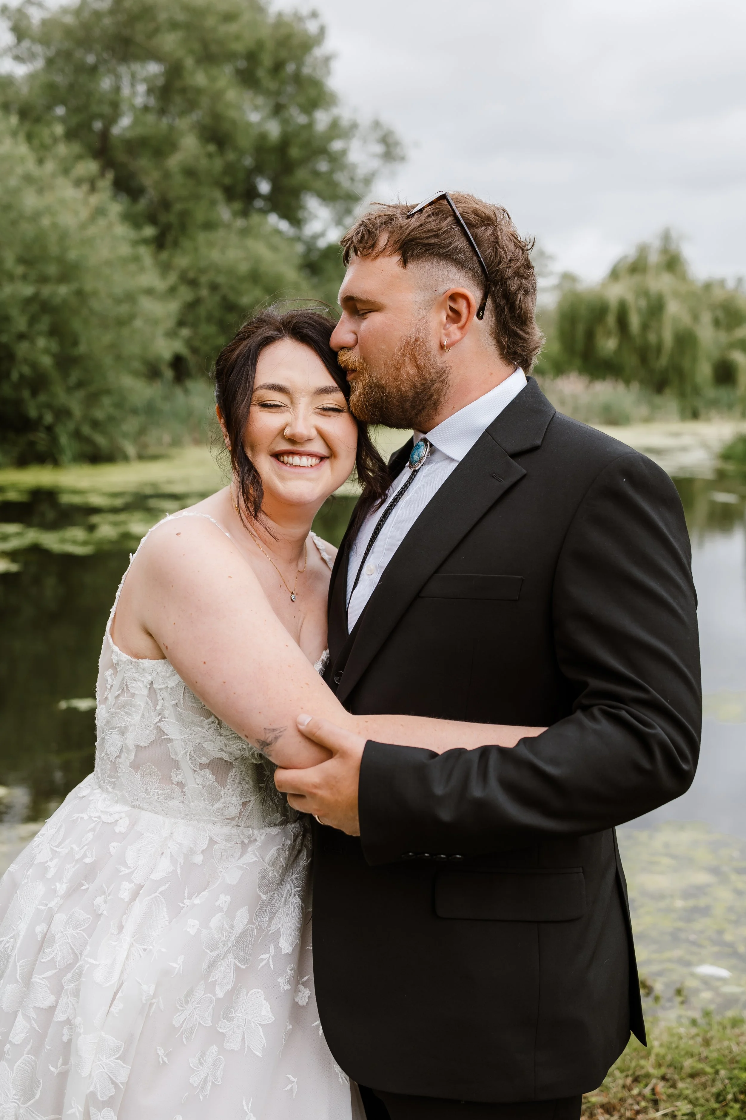 A smiling bride and a groom are embracing near a pond with green trees behind them, outdoors on a cloudy day.