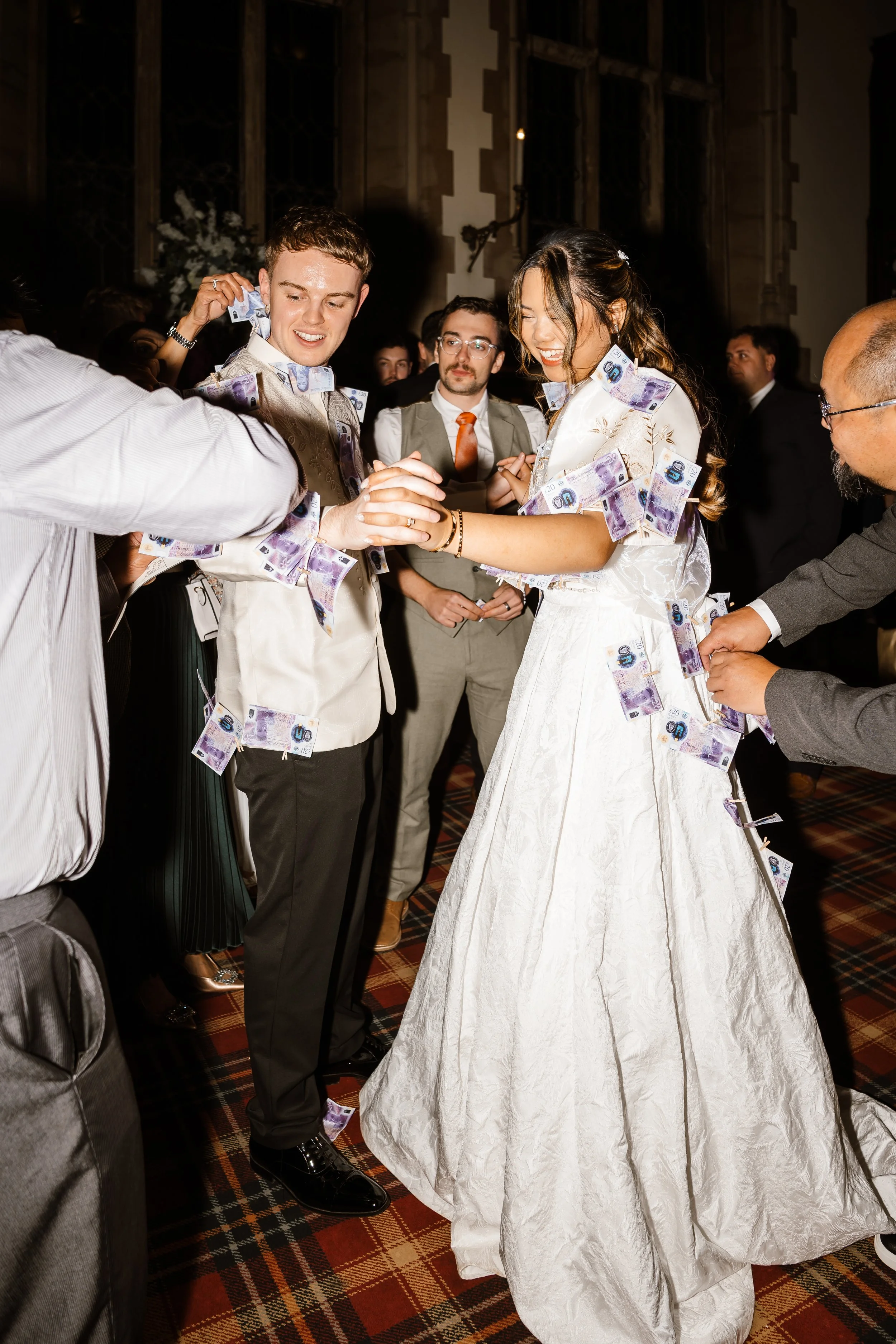 People celebrating at a wedding, with some throwing or wearing $50 bills, in a dimly lit room with a patterned carpet, and ornate wooden walls.