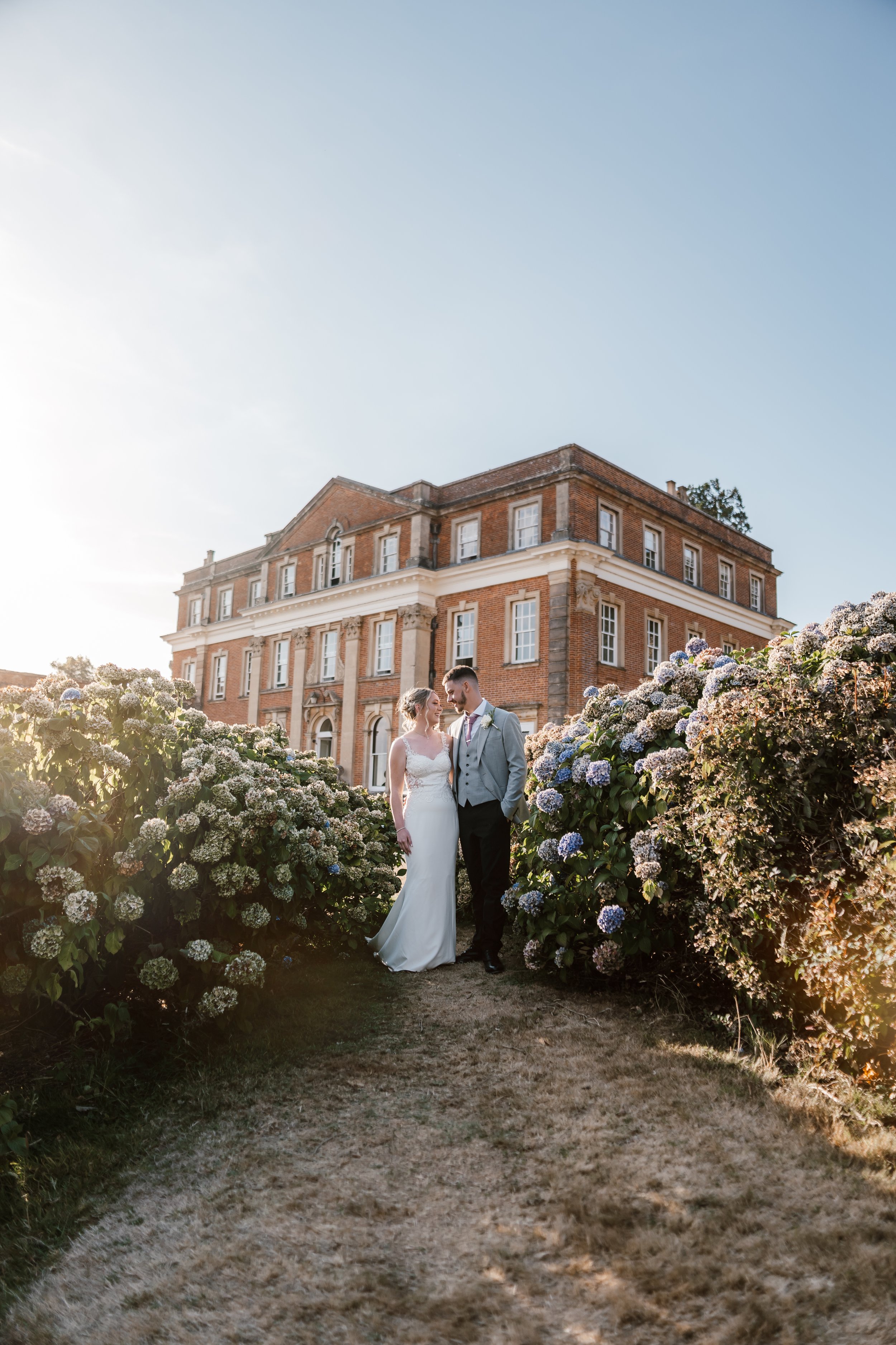 Bride and groom standing together outdoors near blooming hydrangea bushes, with a historic brick building in the background, during sunset.