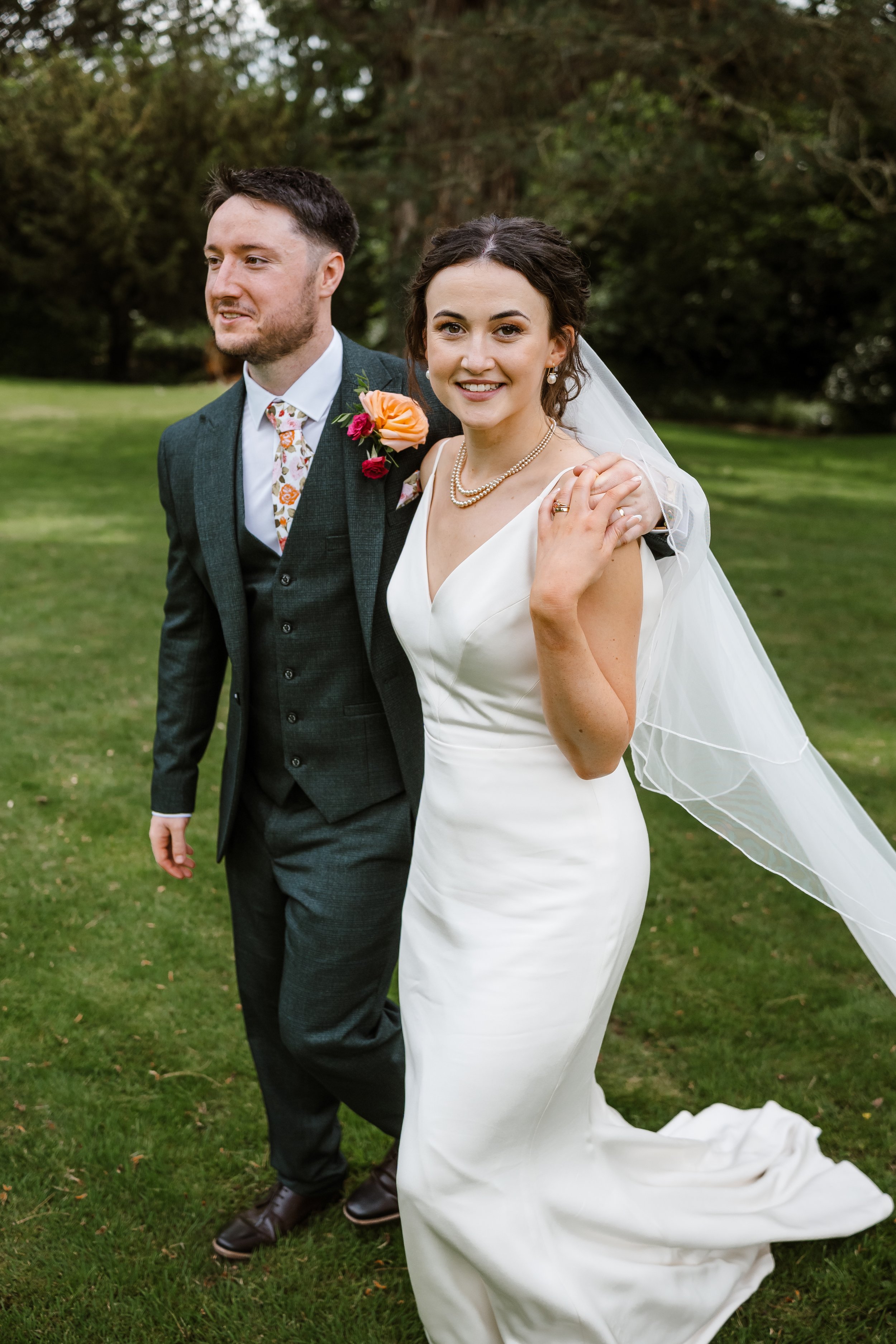 A newlywed couple celebrates outdoors on a grassy area with trees in the background. The bride wears a white wedding dress, pearl necklace, pearl earrings, and a veil. The groom, wearing a gray suit with a colorful floral tie and a boutonniere with o