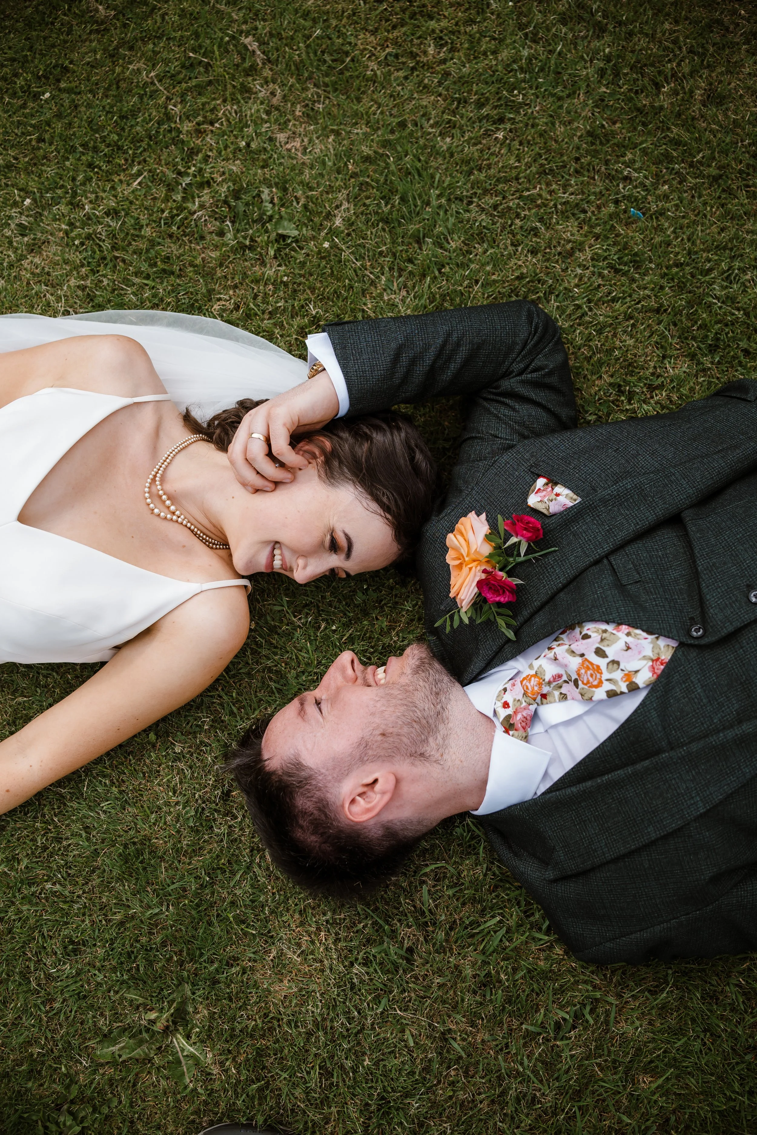 A smiling woman in a white dress and pearl necklace lying on grass next to a man in a suit with a floral shirt and boutonniere. Both are lying on their sides, facing each other, with their heads close together.