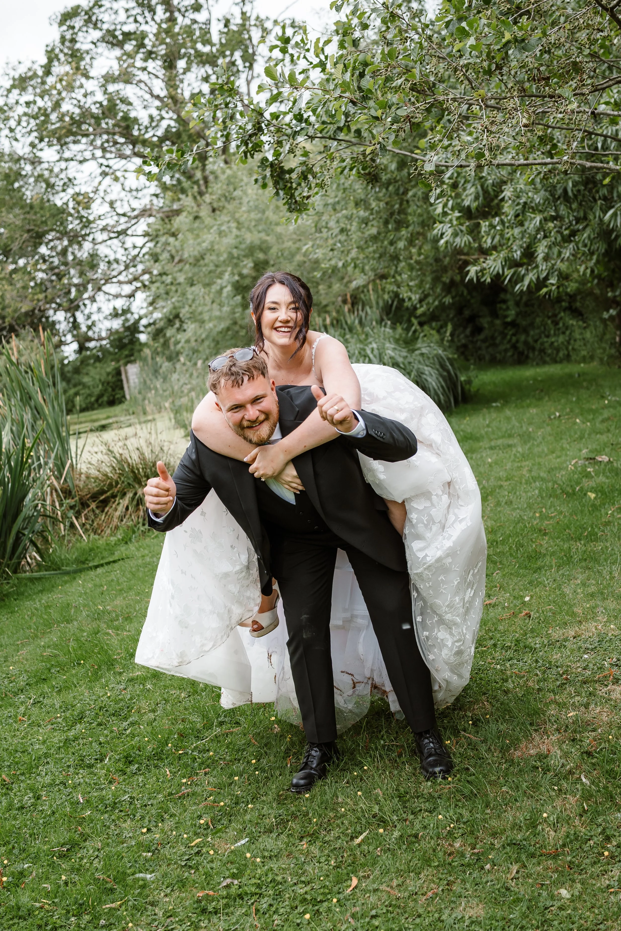 A newly married couple in wedding attire outdoors, with the man carrying the woman on his back. They are smiling and appear joyful, standing on a lush green lawn with trees and bushes in the background.