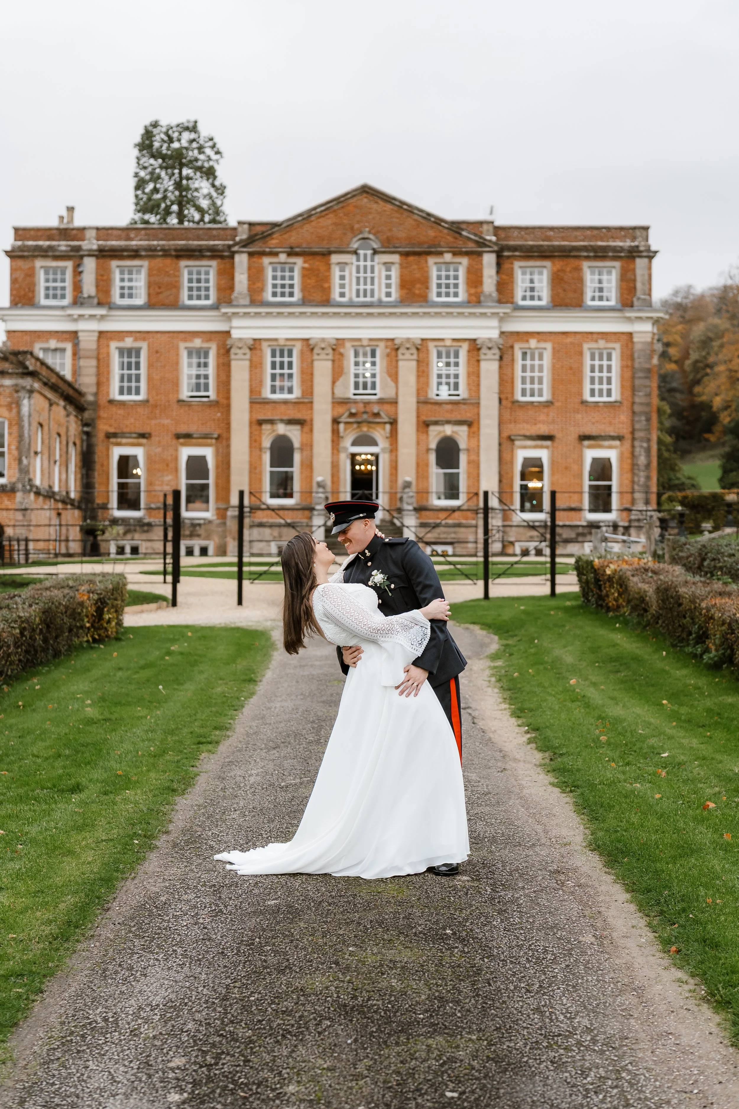 A bride and groom in wedding attire sharing a dance outdoors in front of a historic brick mansion with a gravel pathway and manicured green lawns.